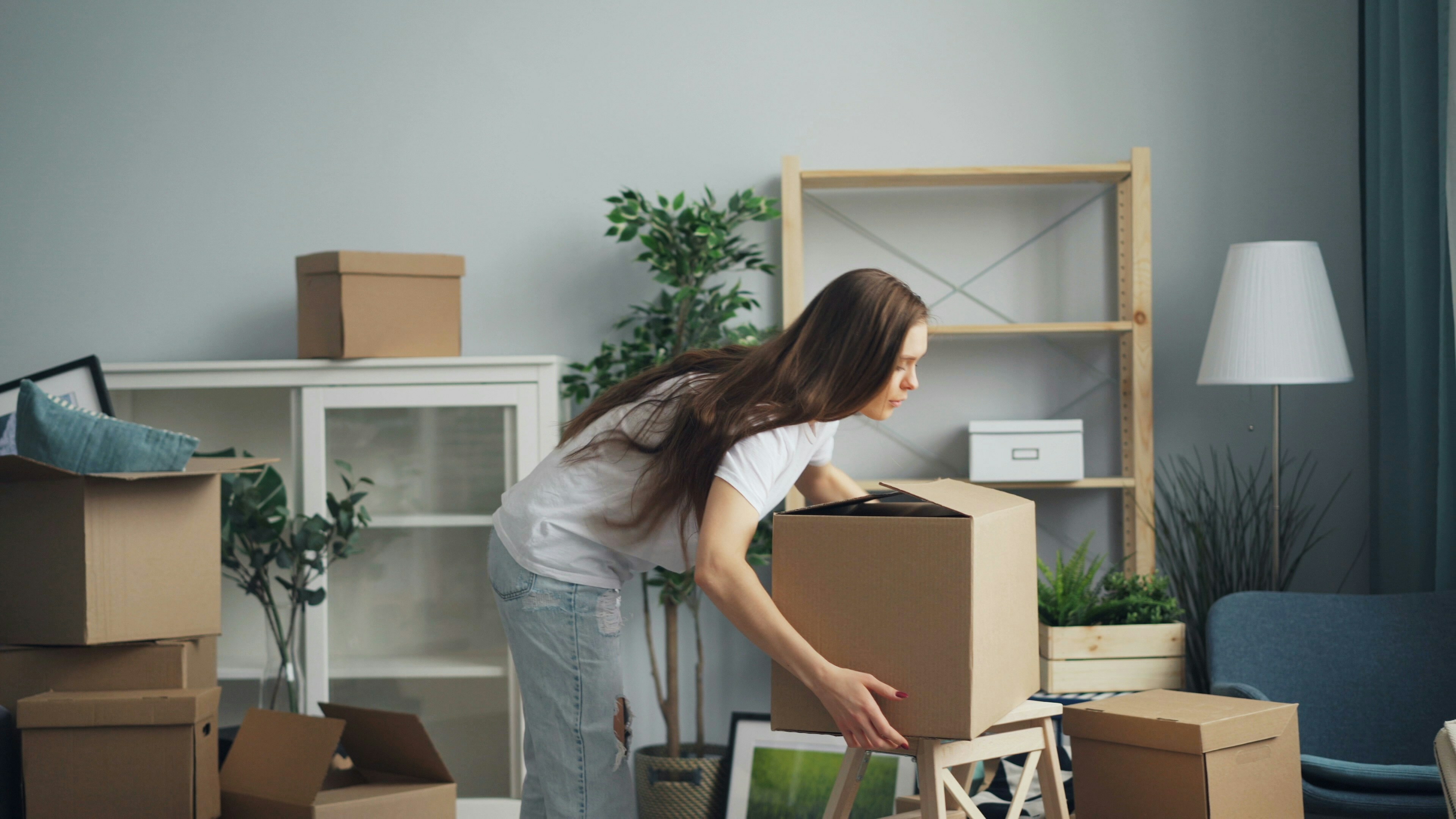 Modern apartment interior showcasing a tidy space with a young woman unpacking boxes, surrounded by stylish d&eacute;cor and greenery.