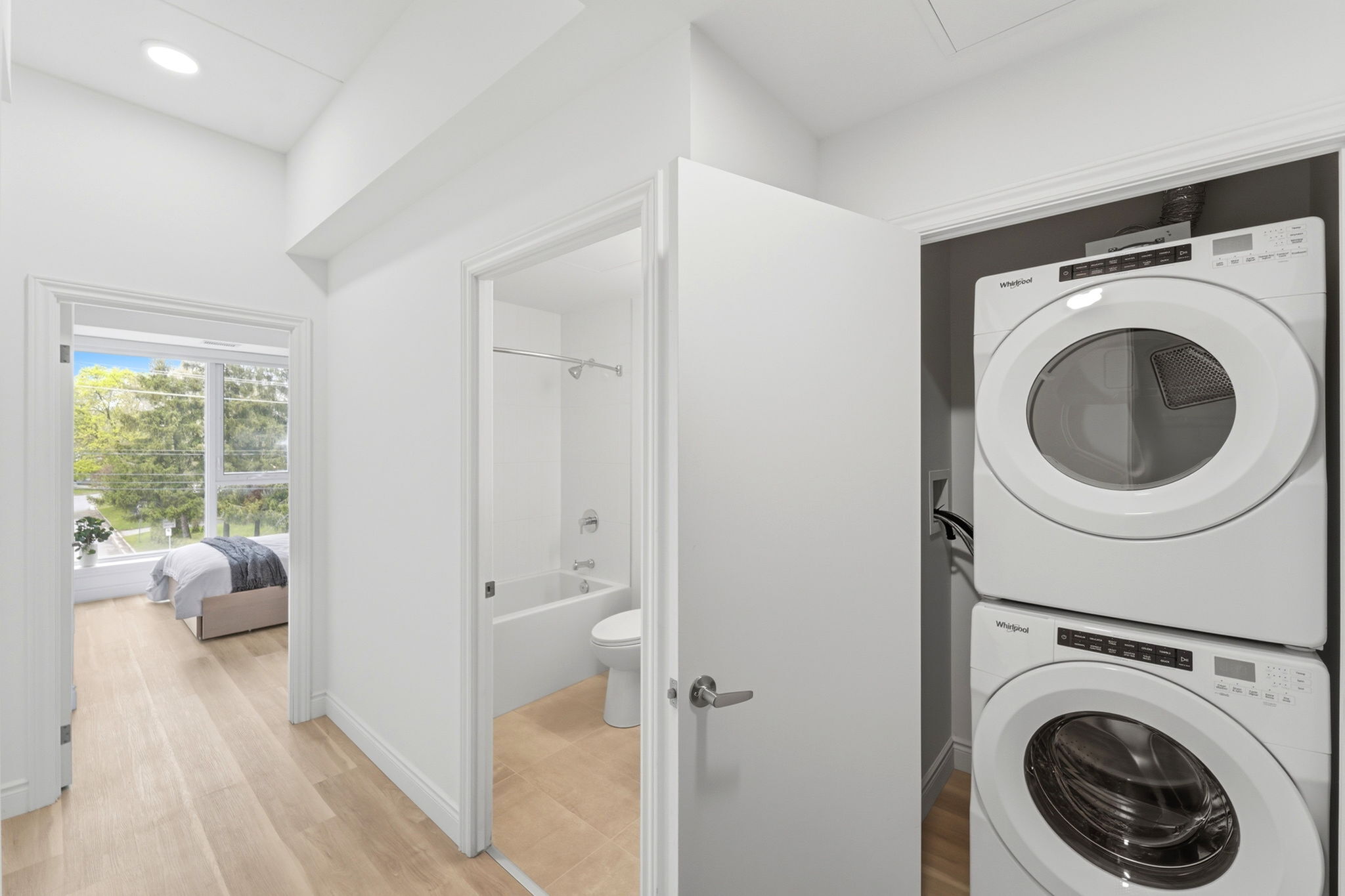 Contemporary apartment hallway featuring a convenient in-unit laundry with stacked washer and dryer, leading to a bright bedroom with large windows and a modern bathroom.
