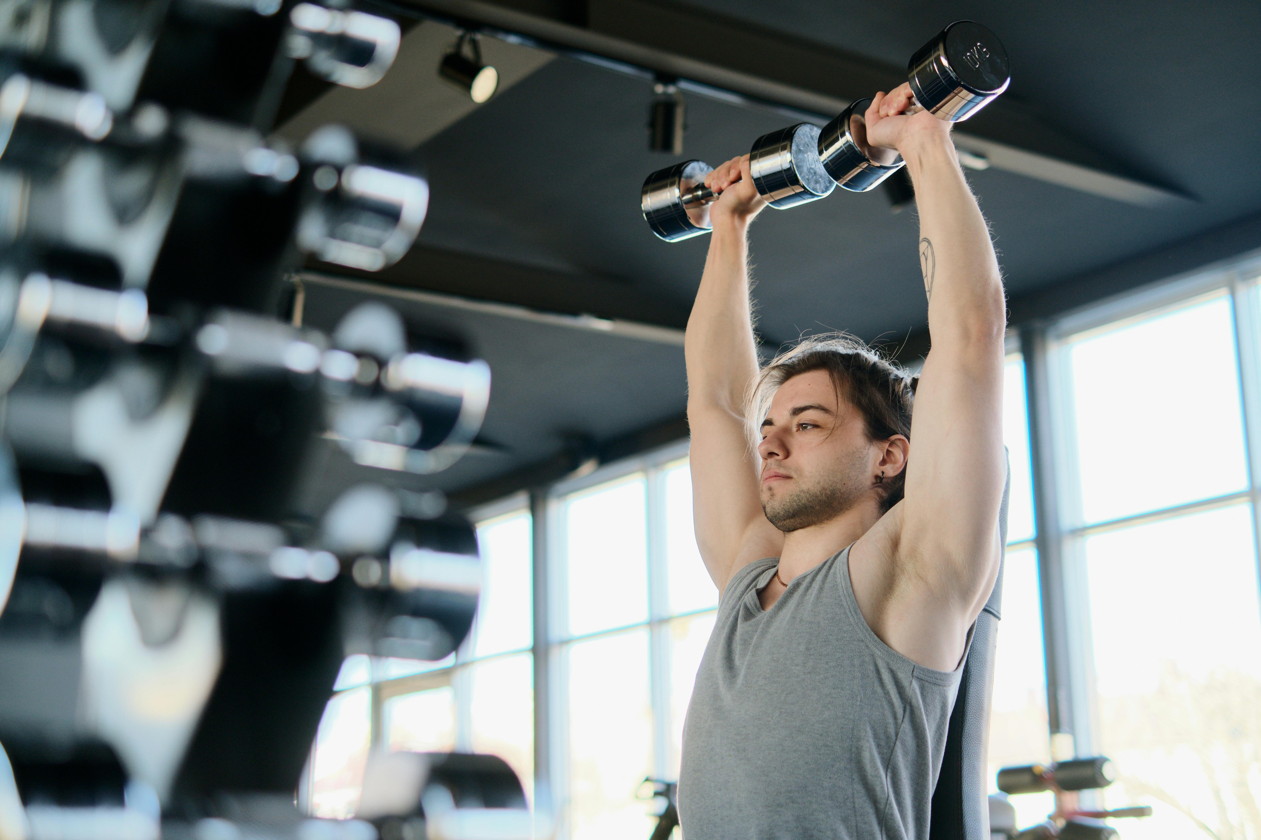 Focused individual lifting dumbbells in a modern, well-lit gym with large windows and sleek equipment.
