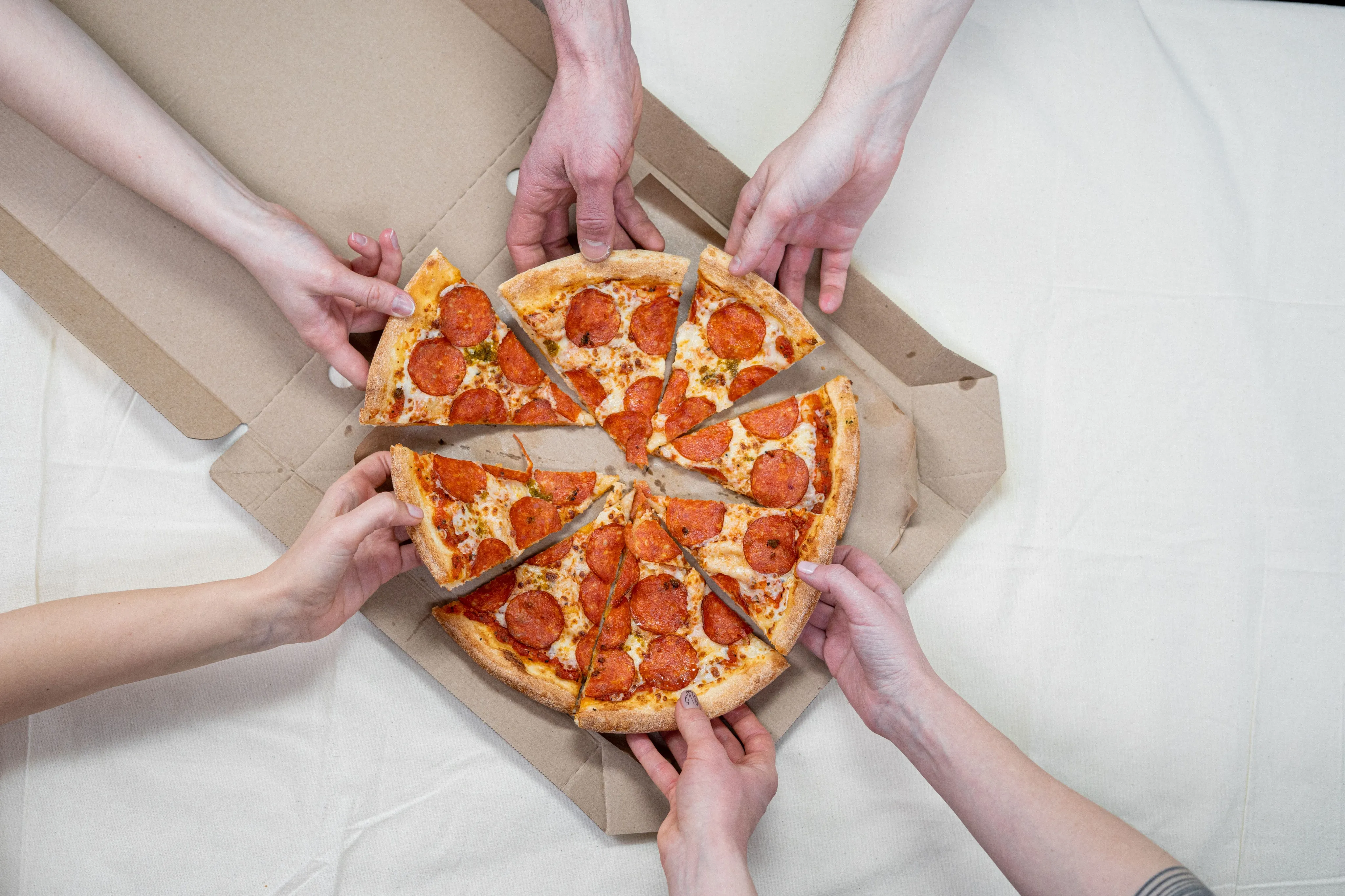 Top view of multiple hands eagerly reaching for delicious pepperoni pizza slices in an open box, emphasizing a shared and appetizing experience.