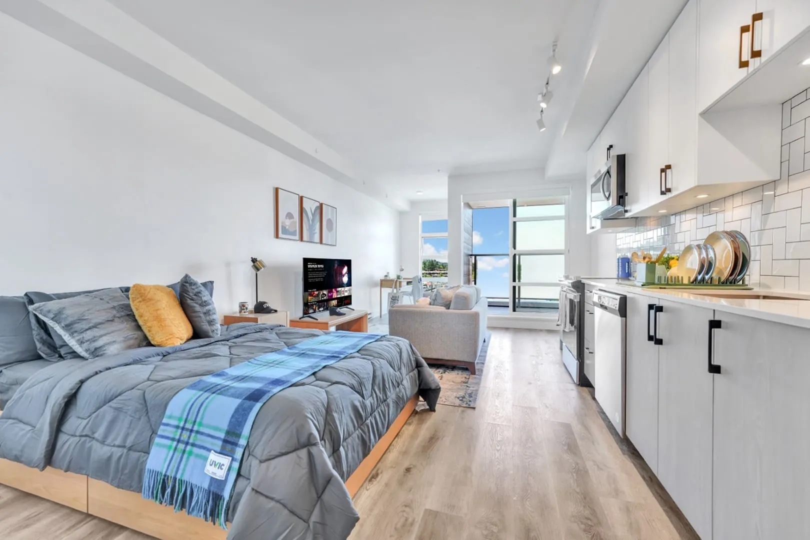 Bright and modern studio apartment featuring a cozy bed, stylish sofa, sleek kitchen, and a window with stunning sky views.