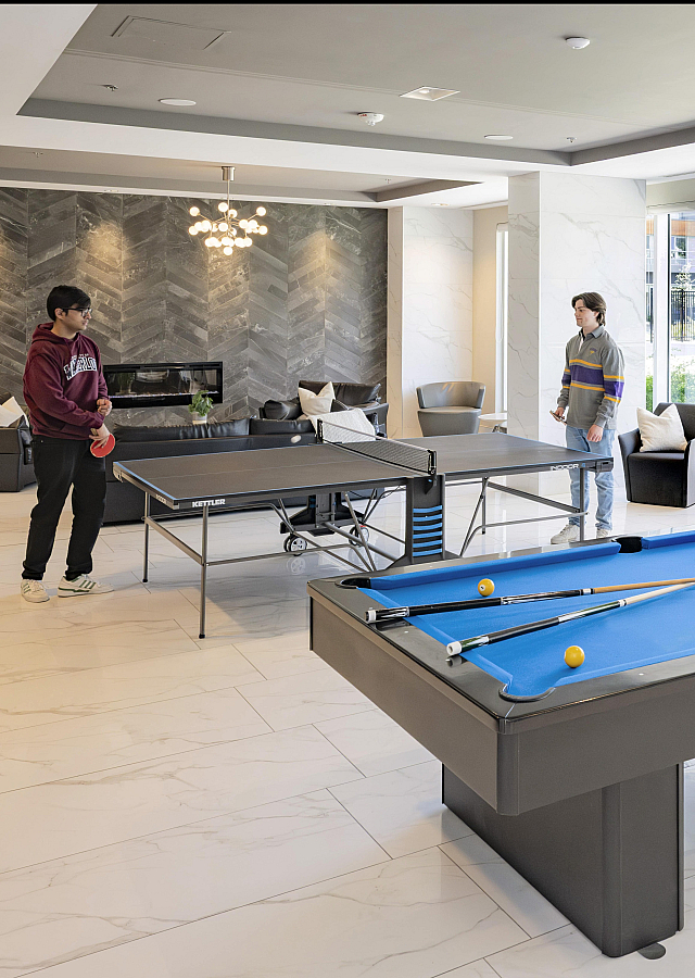 Modern recreation room with a pool table, ping pong setup, and cozy seating area, accented by large windows and elegant lighting.