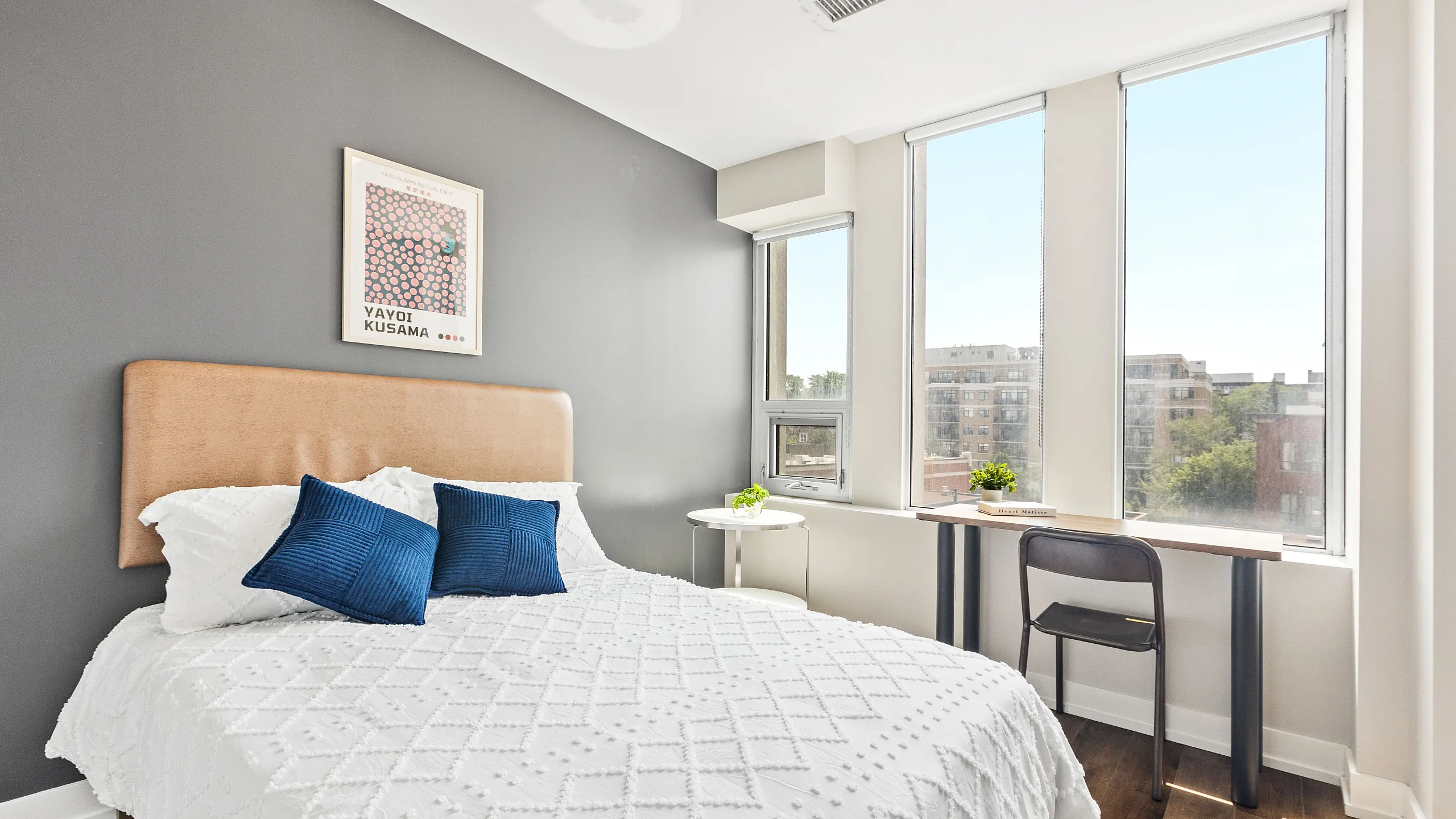Chic bedroom with modern decor, featuring a plush bed adorned with blue accent pillows, large windows for natural light, and a stylish desk area.