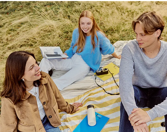 Vibrant picnic scene with three friends enjoying a sunny day on a striped blanket in a grassy field, surrounded by books and a vintage camera.