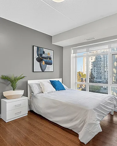 Bright and modern bedroom with hardwood floors, featuring a large window view, stylish artwork, and a cozy bed with accent pillows.