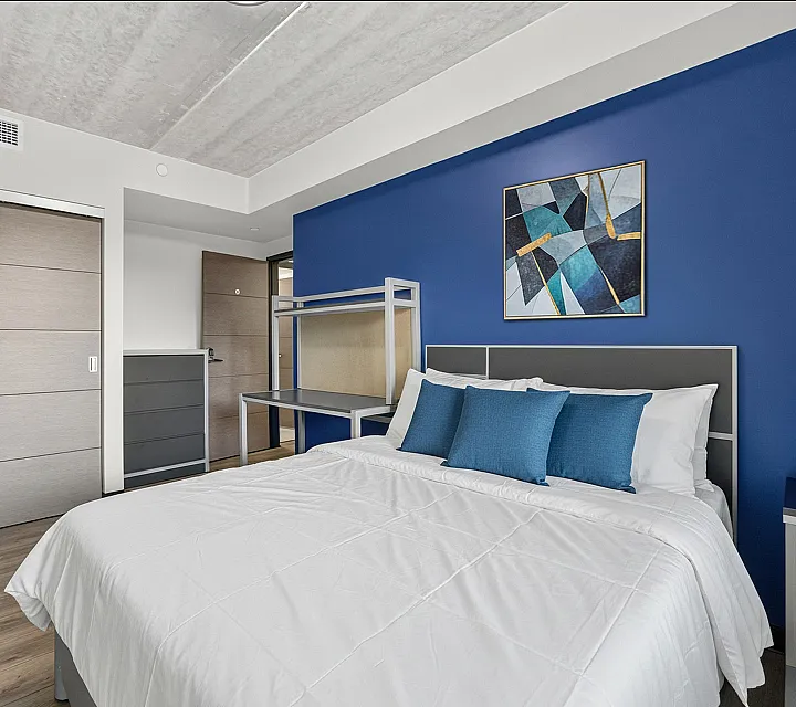 Stylish bedroom with modern decor, featuring a plush bed adorned with blue accent pillows, abstract artwork on a vibrant blue wall, and sleek furnishings for a contemporary feel.