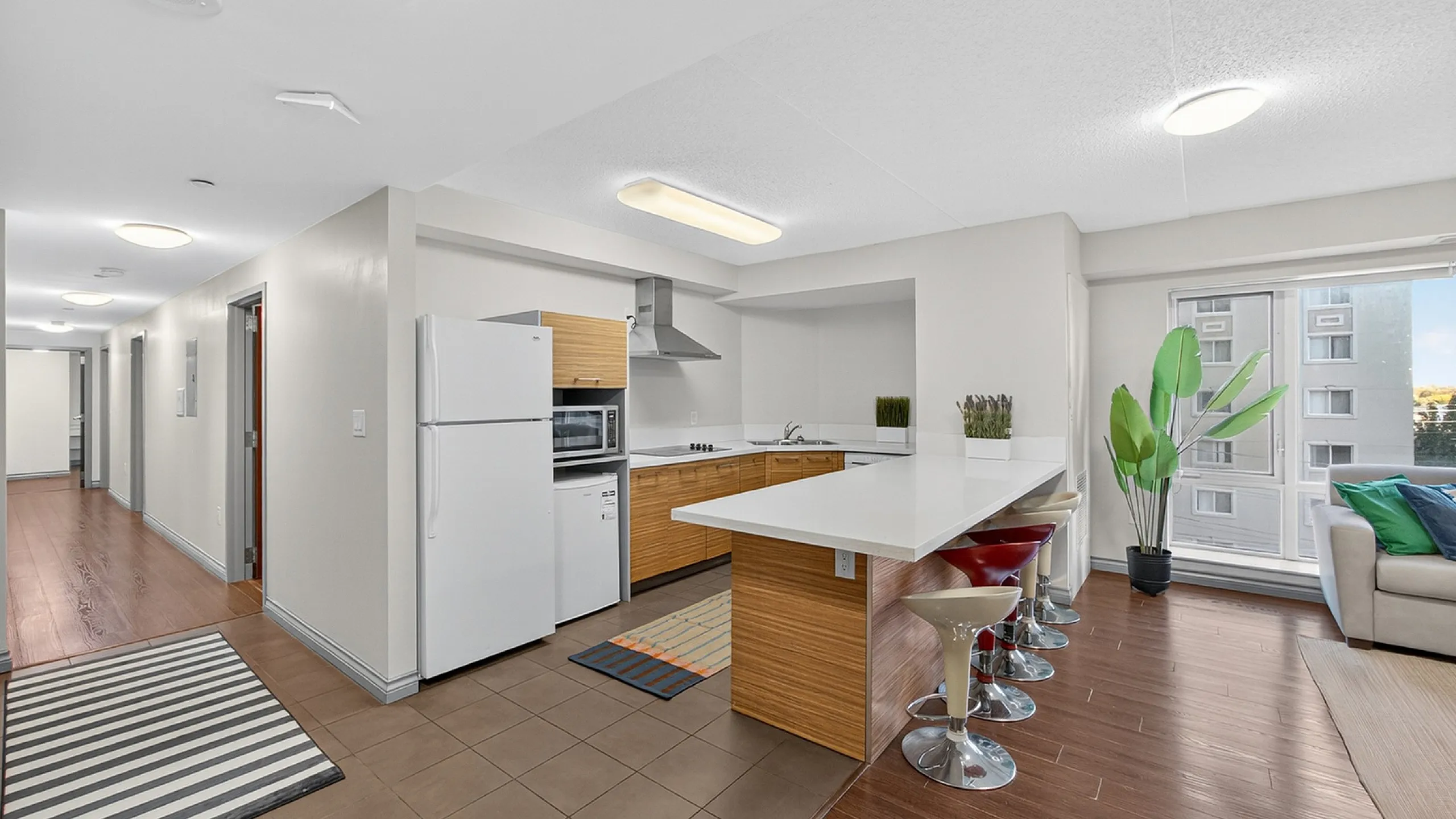 Modern open-concept kitchen with sleek white countertops, cozy bar seating, and abundant natural light, adjacent to a stylish living area with large windows.