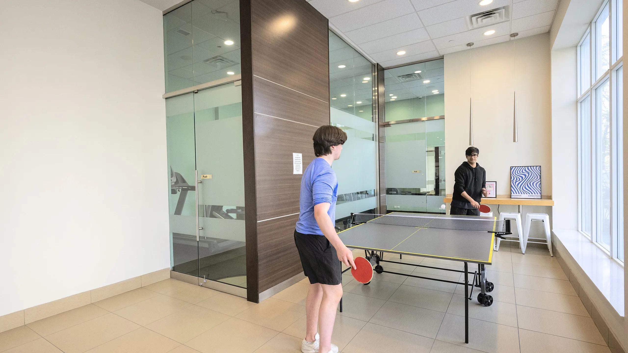 Modern recreational area with a sleek table tennis setup, natural light pouring in from large windows, and a glass-walled exercise room in the background.
