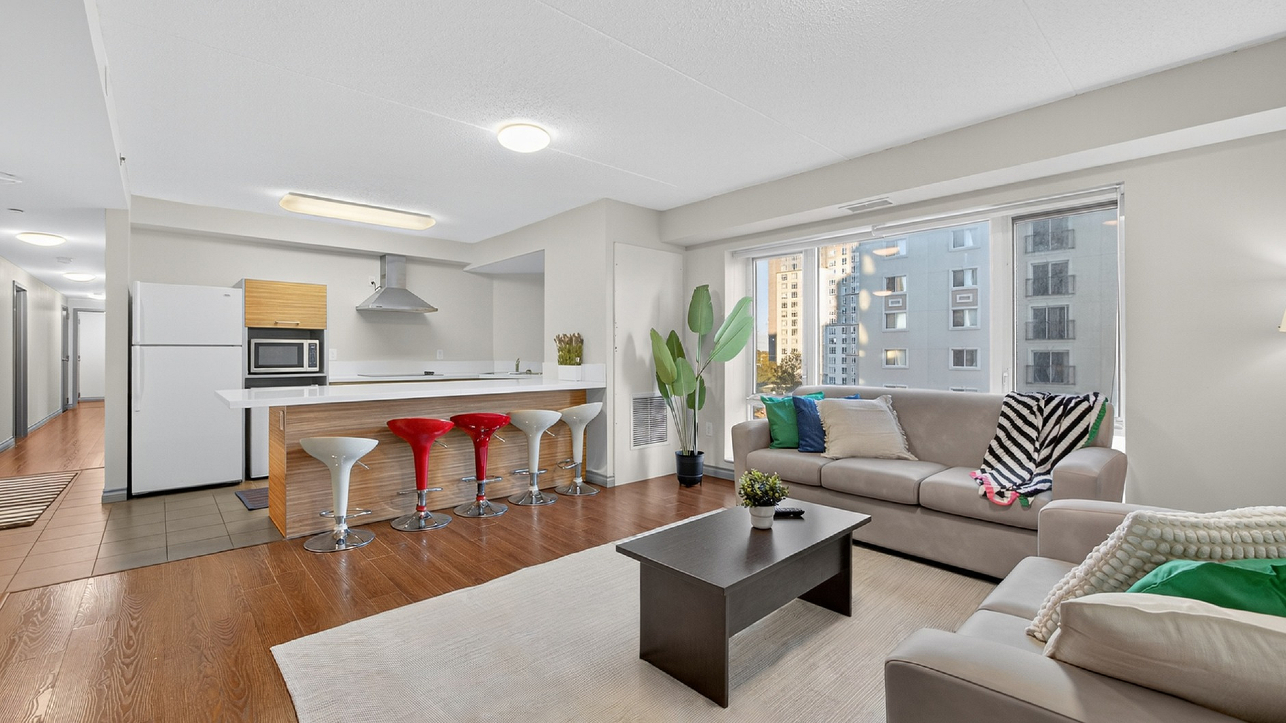 Modern open-plan living area featuring a stylish kitchen with vibrant bar stools, a cozy seating space with plush sofas, and large windows offering stunning city views.