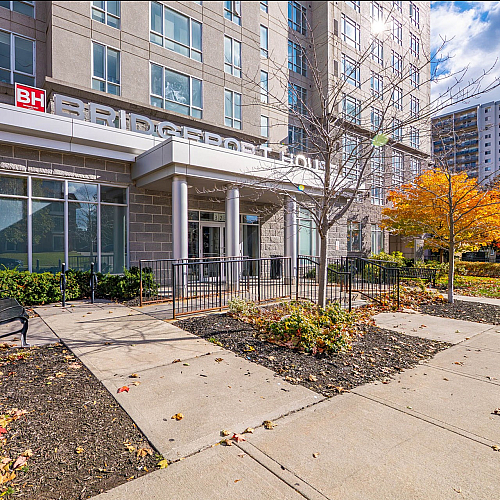 Modern apartment building entrance with large windows and a welcoming pathway lined with colorful autumn foliage.