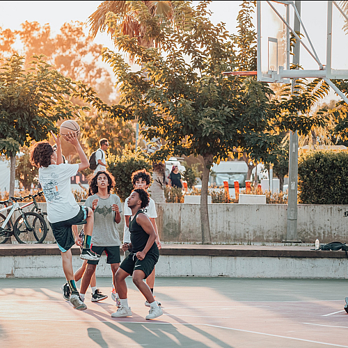 Dynamic outdoor basketball game in progress at sunset with players actively engaged, surrounded by lush greenery and a vibrant atmosphere.