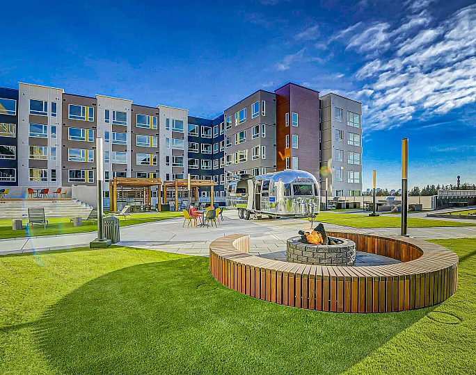 Vibrant outdoor space of a modern apartment complex featuring stylish seating, a sleek Airstream trailer, and an inviting fire pit under a clear blue sky.