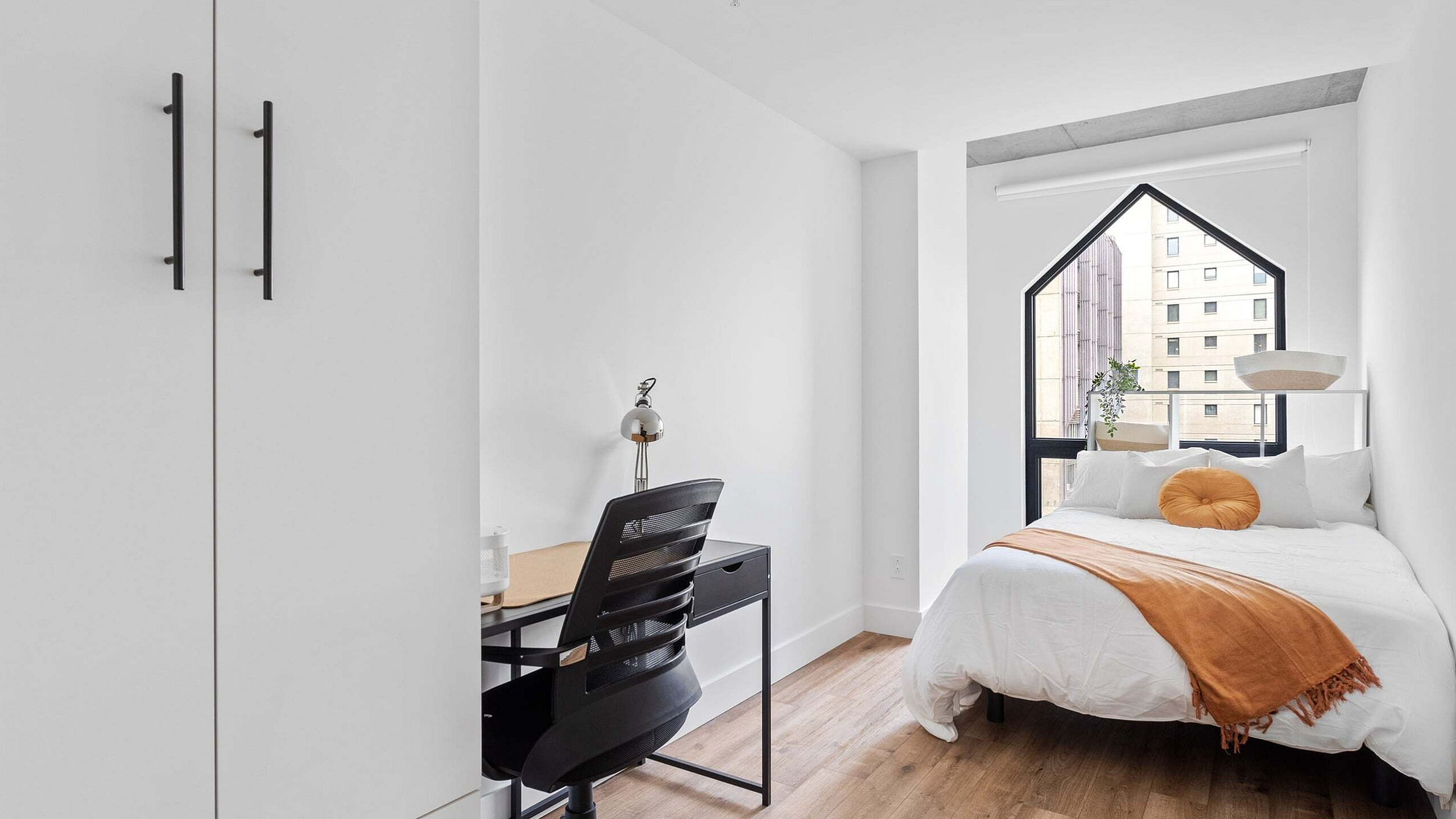Bright and modern bedroom with sleek furniture, featuring a cozy bed with orange accents, a stylish desk setup, and an arched window offering a city view.