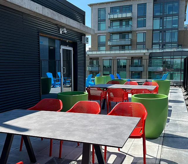 Chic rooftop patio with vibrant red chairs and modern tables, overlooking a stylish apartment building under a clear blue sky.