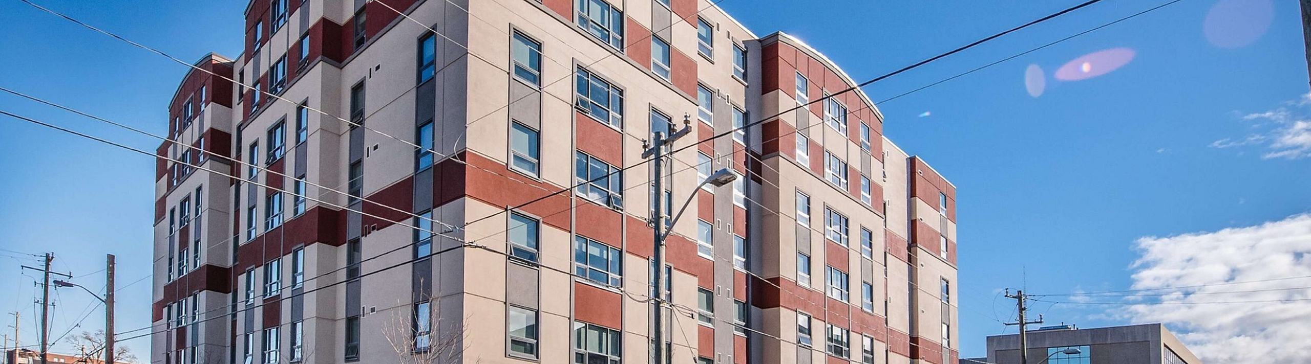 Modern apartment building with a red and beige facade, large windows, and urban surroundings under a clear blue sky.