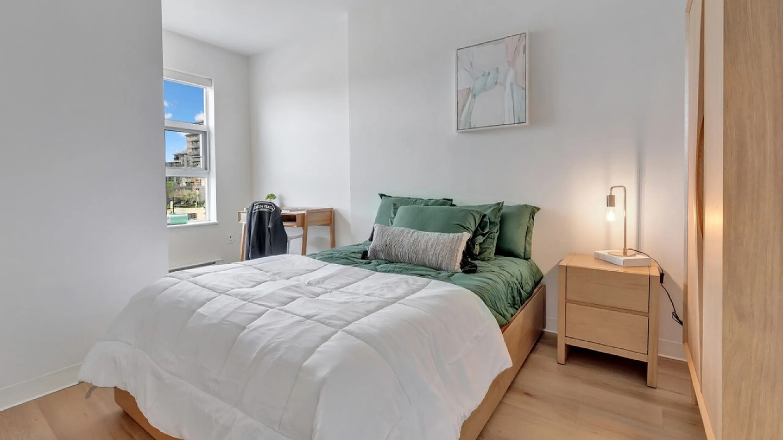 Bright and cozy bedroom featuring a comfortable bed with green accents, modern nightstand, wooden flooring, and a large window with a charming city view.