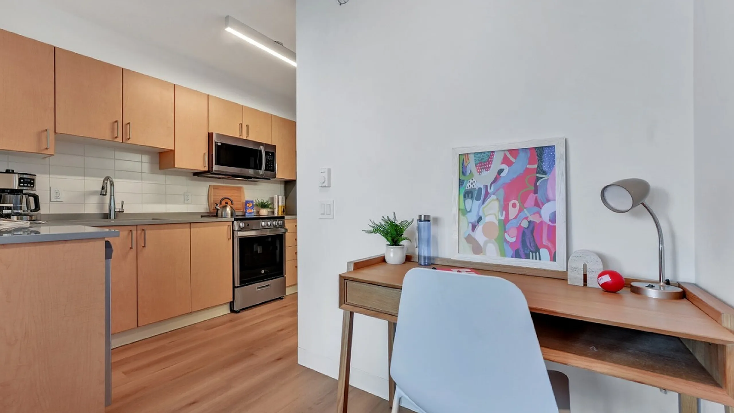 Modern kitchen with sleek wooden cabinets, stainless steel appliances, and a cozy desk area with colorful artwork.