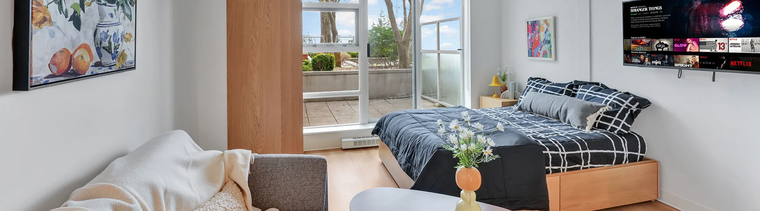 Chic studio apartment featuring a cozy bed, stylish decor, and a large window opening to a sunny patio.