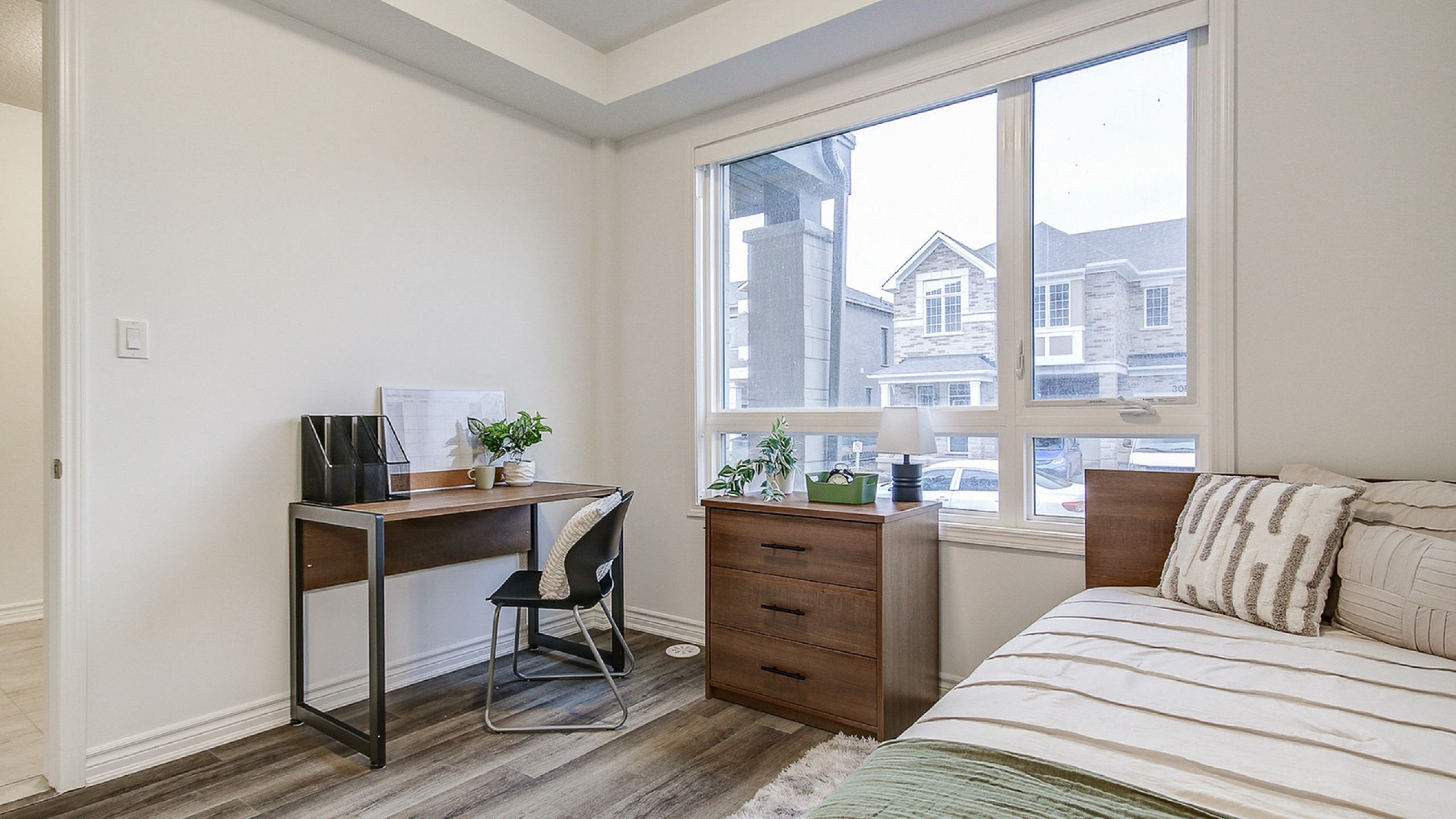 Bright and inviting bedroom featuring a cozy work desk by a large window, showcasing charming neighborhood views and elegant wood flooring.