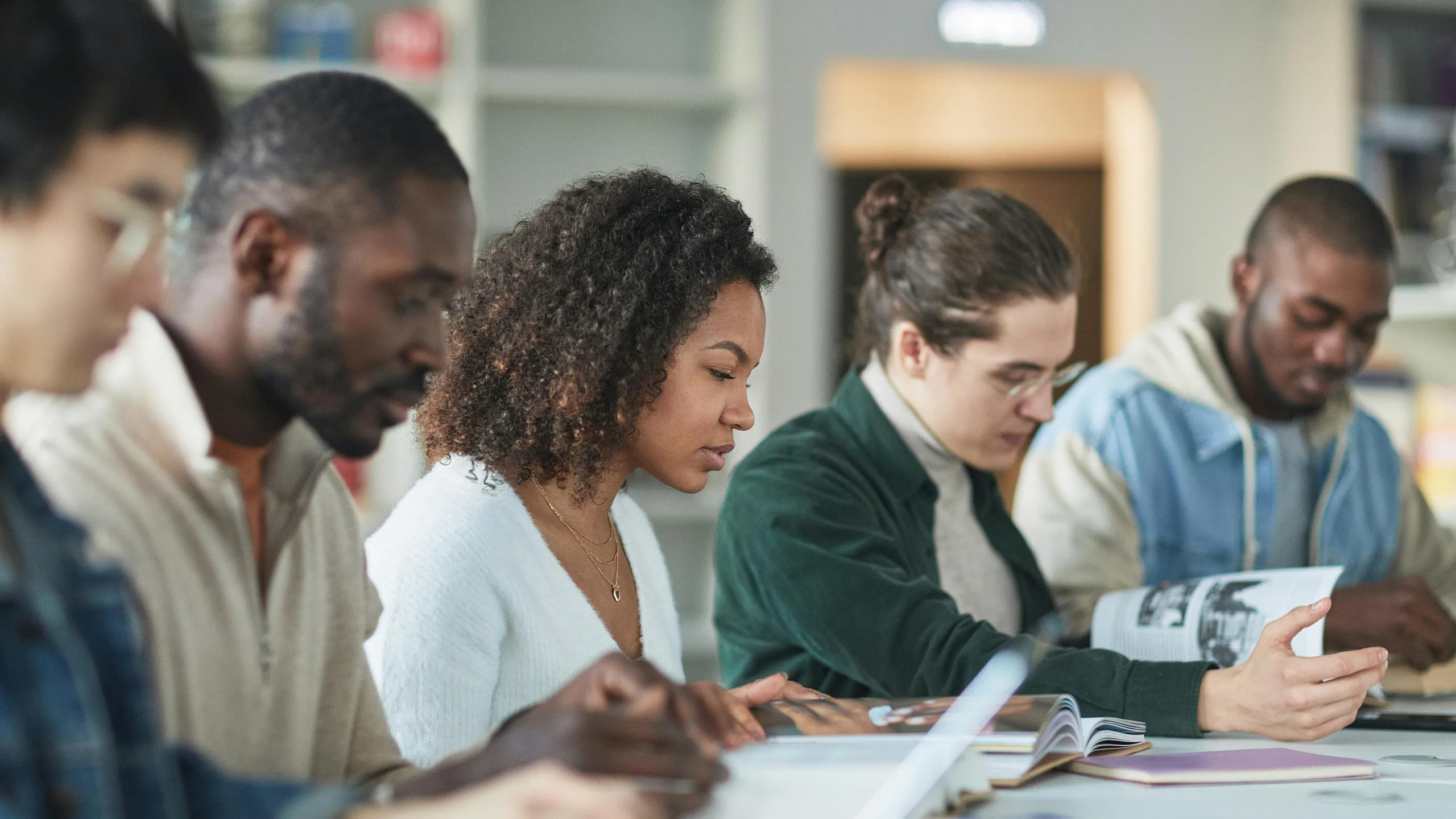 Diverse group of students engaged in focused study at a library table, surrounded by open books and natural light.