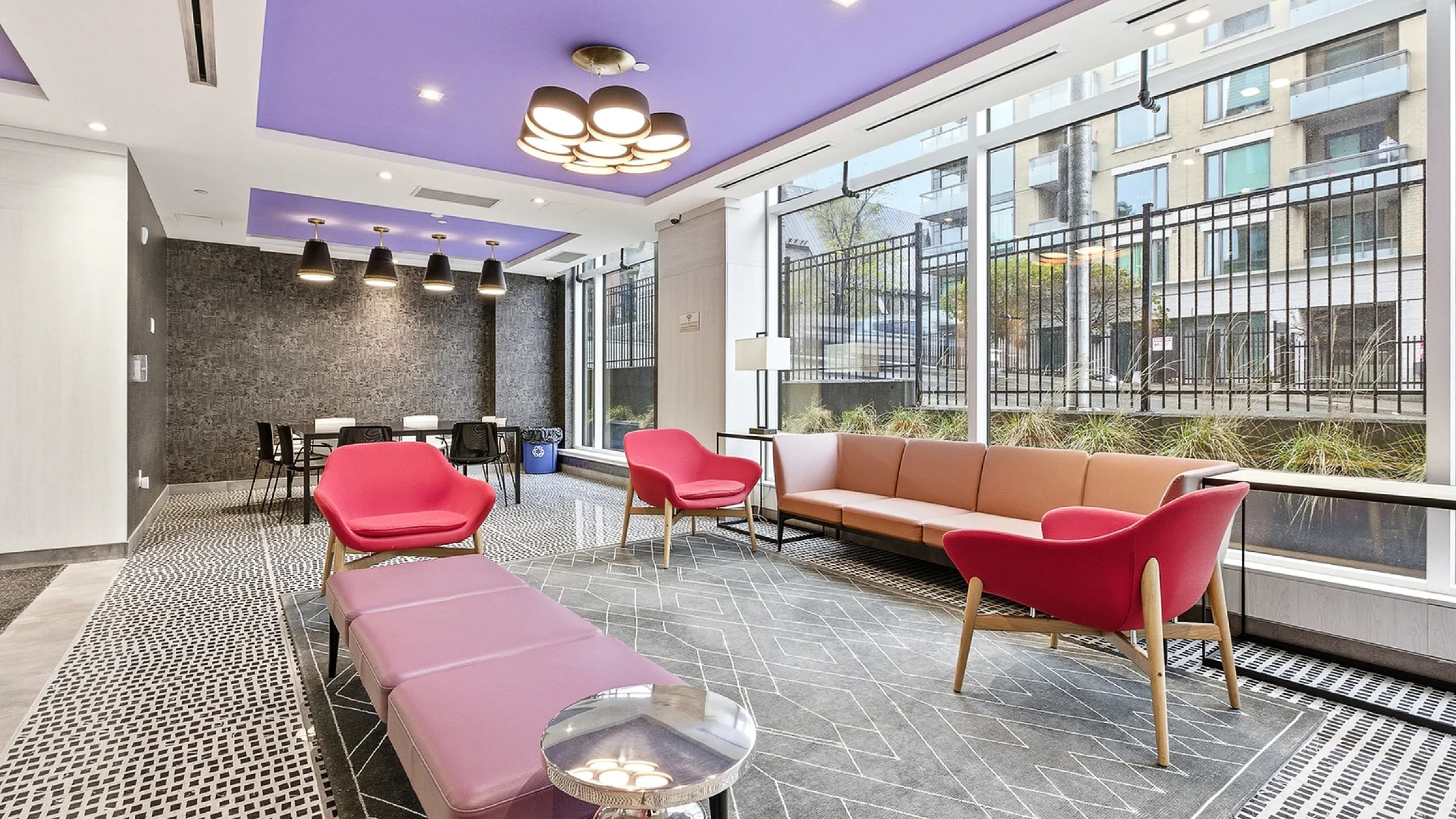 Chic modern lounge area with vibrant purple ceiling, stylish lighting fixtures, cozy pink and red seating, and large windows offering a city view.