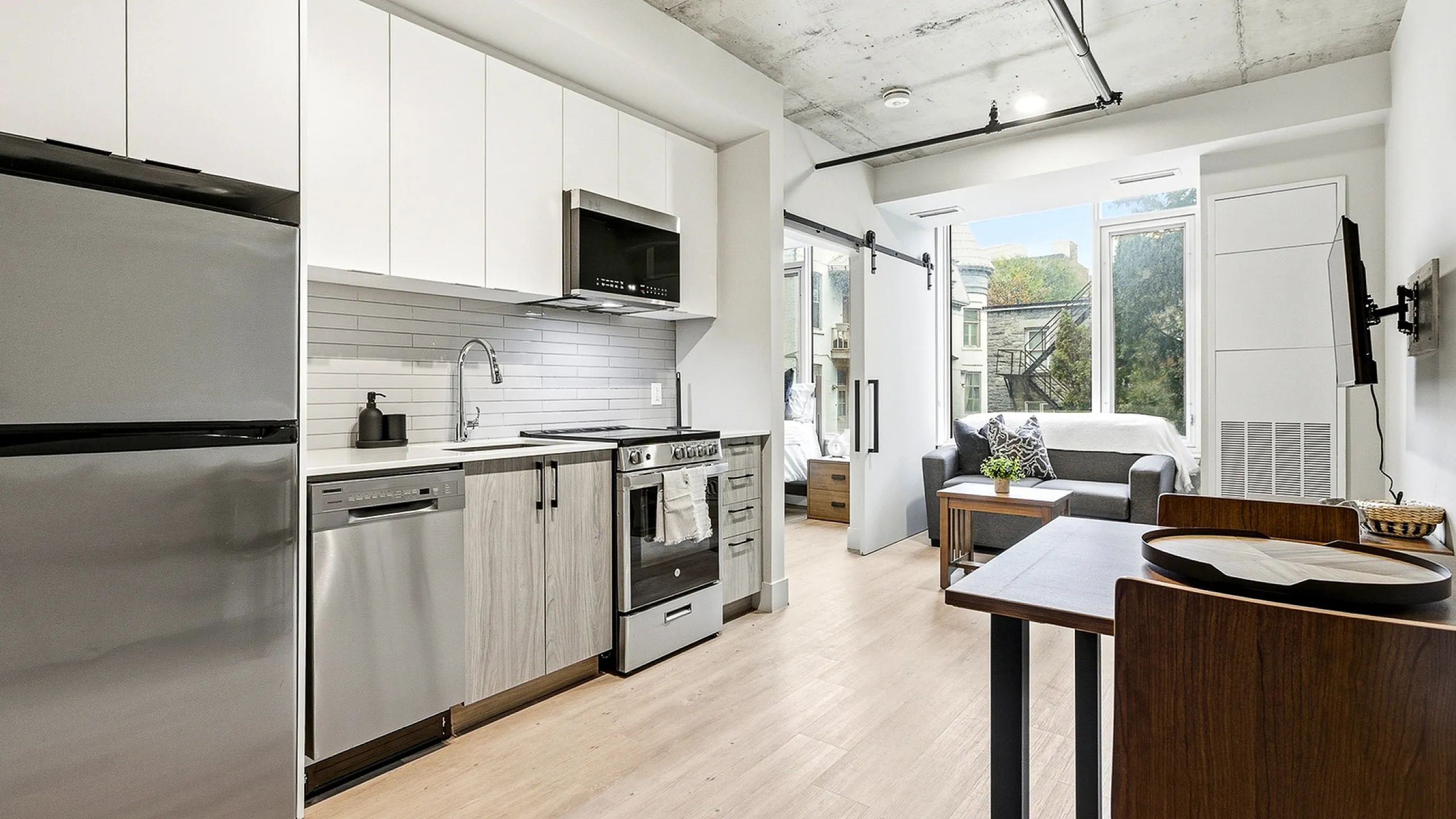 Modern apartment kitchen and living space featuring sleek stainless steel appliances, contemporary cabinetry, and a cozy seating area with large windows.
