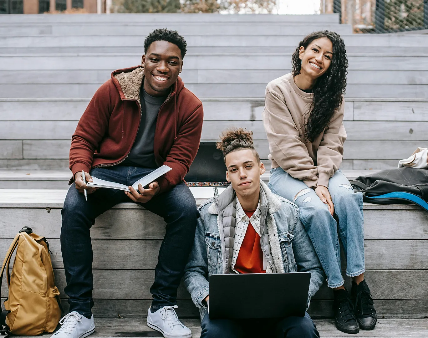 Three friends sitting on outdoor steps, smiling and enjoying a sunny day with backpacks and a laptop.
