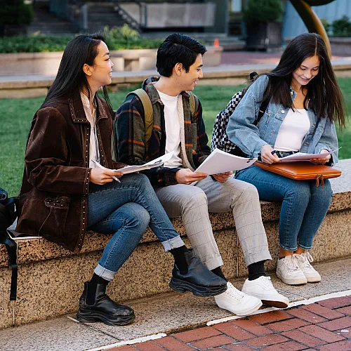 Three students sit on a bench, smiling and engaged in conversation, with textbooks and backpacks amidst a serene campus lawn setting.