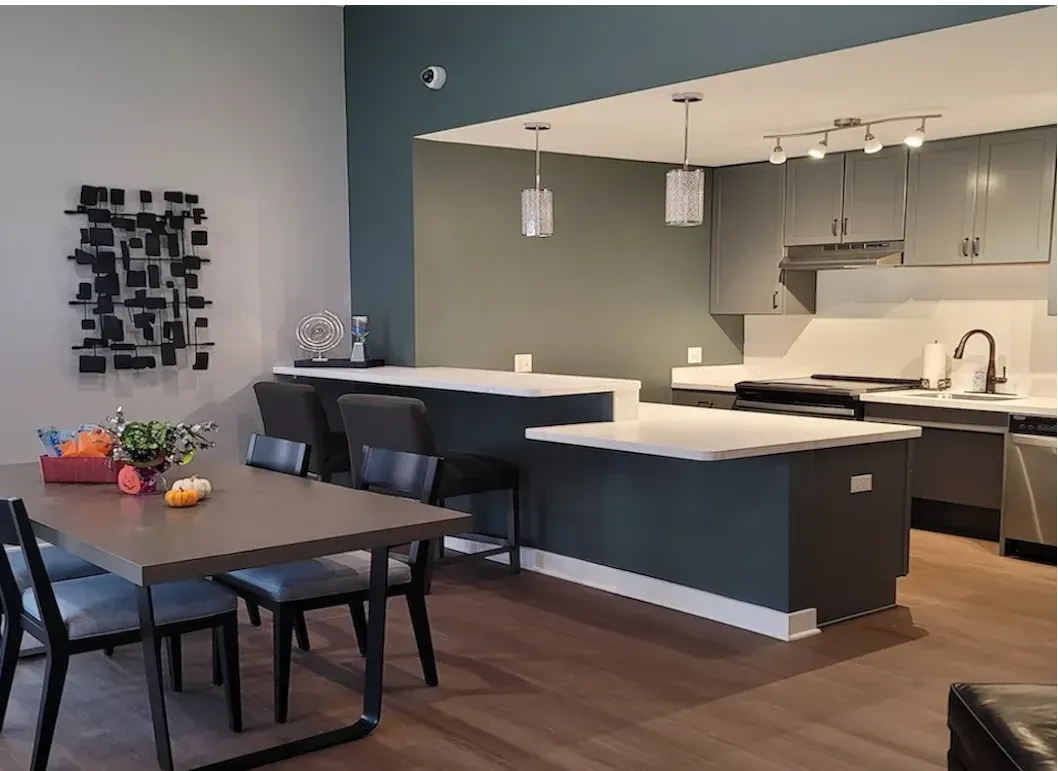 Community Room featuring harwood floors, community ktichen with white quoartz countertops, gray cabinetry. Sage Green walls.  Breakfast bar with two bar stools. Also, a dining room table with four chairs.