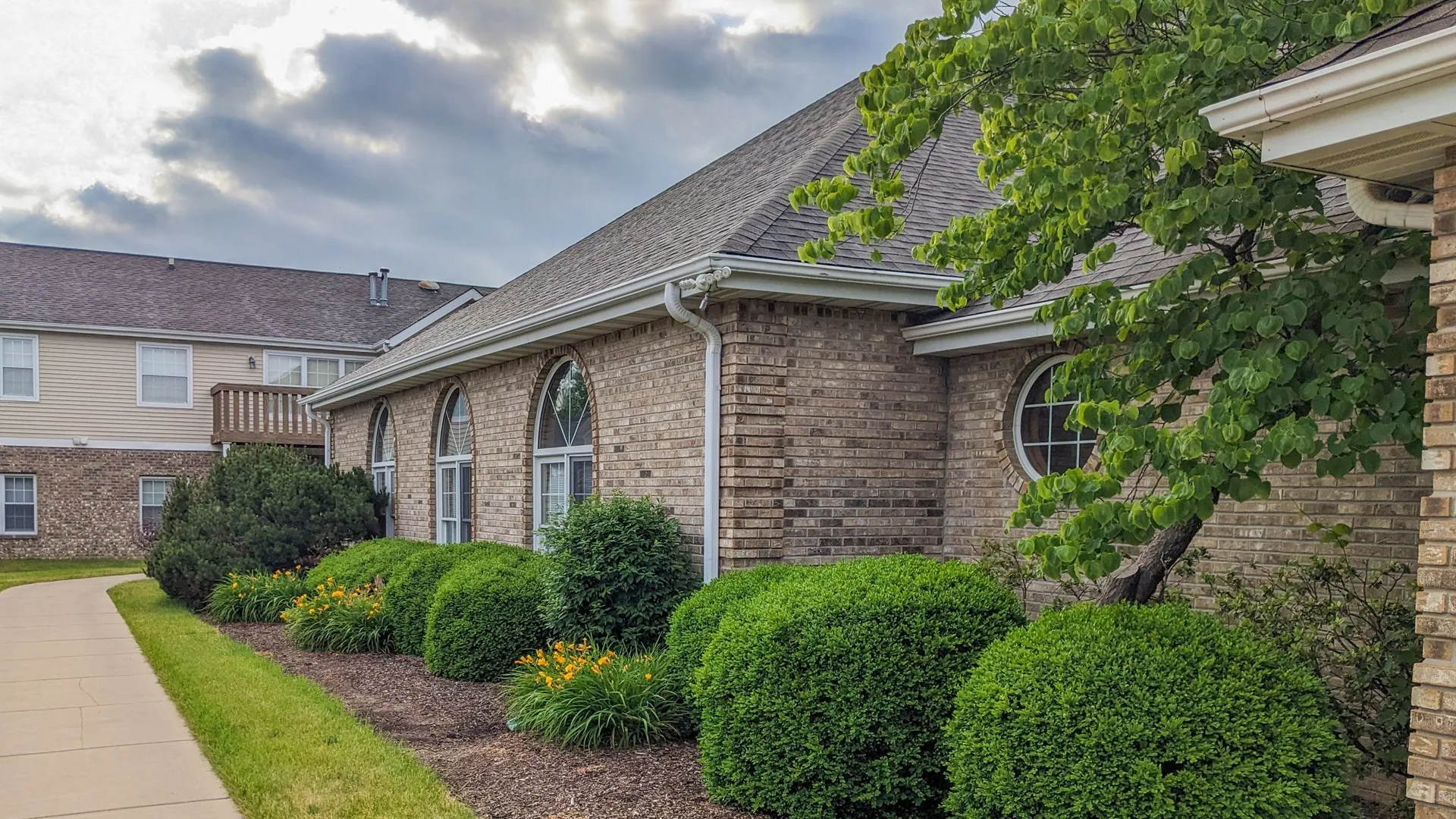 The neutral brick leasing office with arched windows. Behind the office building is an apartment building. It has light tan siding on the top portion of the building, a neutral brick accent on the lower portion of the building, and white tr