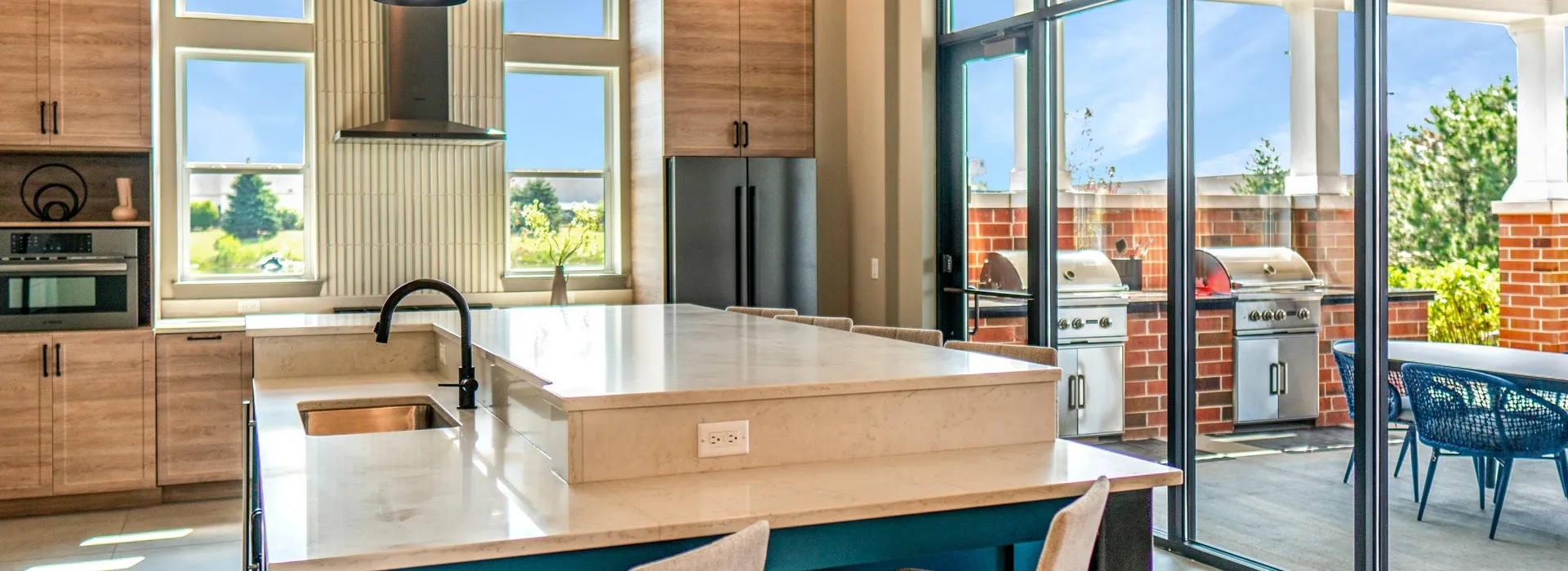 Clubhouse community kitchen with a large island in the middle of the room. Along the back wall is light brown cabinetry, stainless steel appliances, and an off-white tiled middle accent with a large range hood. Windows are on both sides of