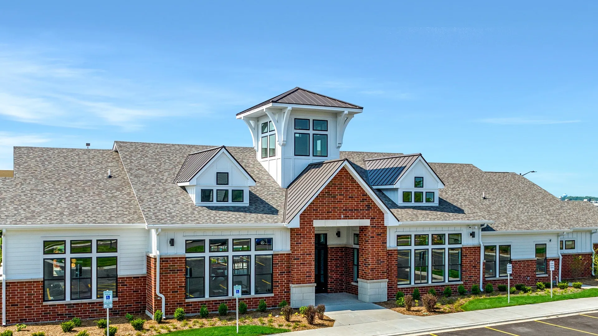 The exterior of the clubhouse with red brick, white siding, and numerous windows to allow natural light to illuminate the inside of the building. In front of the building is landscaping including shrubs, bushes, and grass.