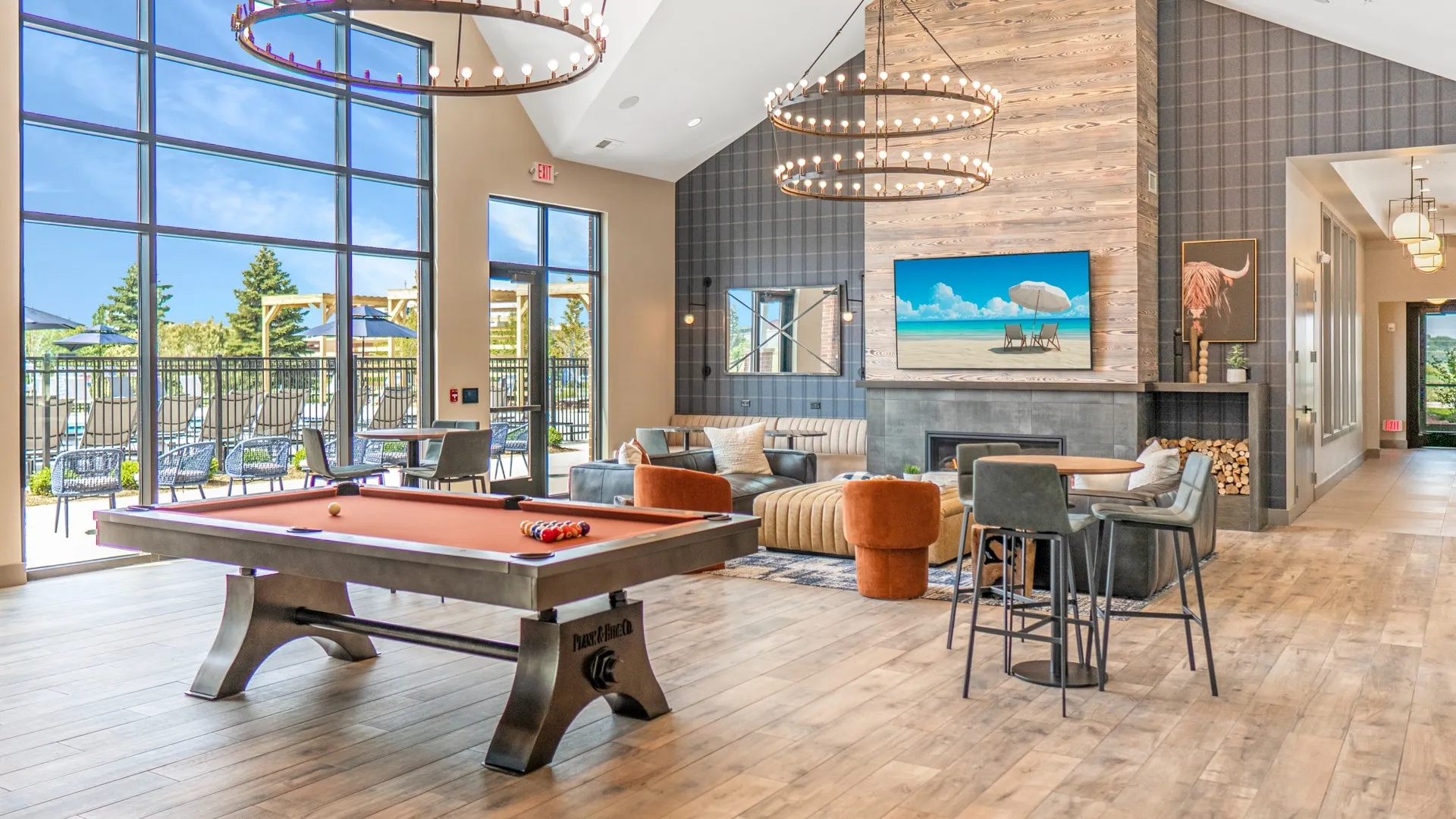 Clubhouse with a large seating area around a fireplace with a TV mounted above. Near the seating area is a pool table with rust-colored felt. The long wall back has floor-to-ceiling windows that look out to the pool area outside.