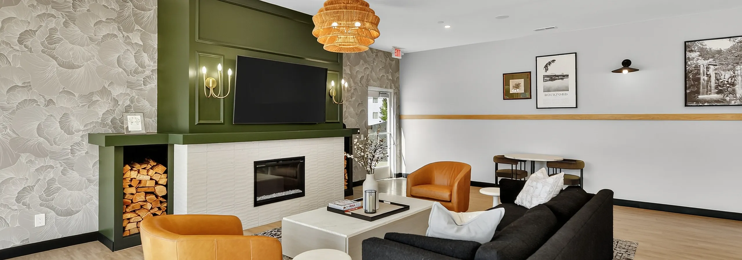 Chic and cozy community room featuring a stylish black sofa, elegant fireplace with wood storage, and warm leather chairs under a decorative pendant light.