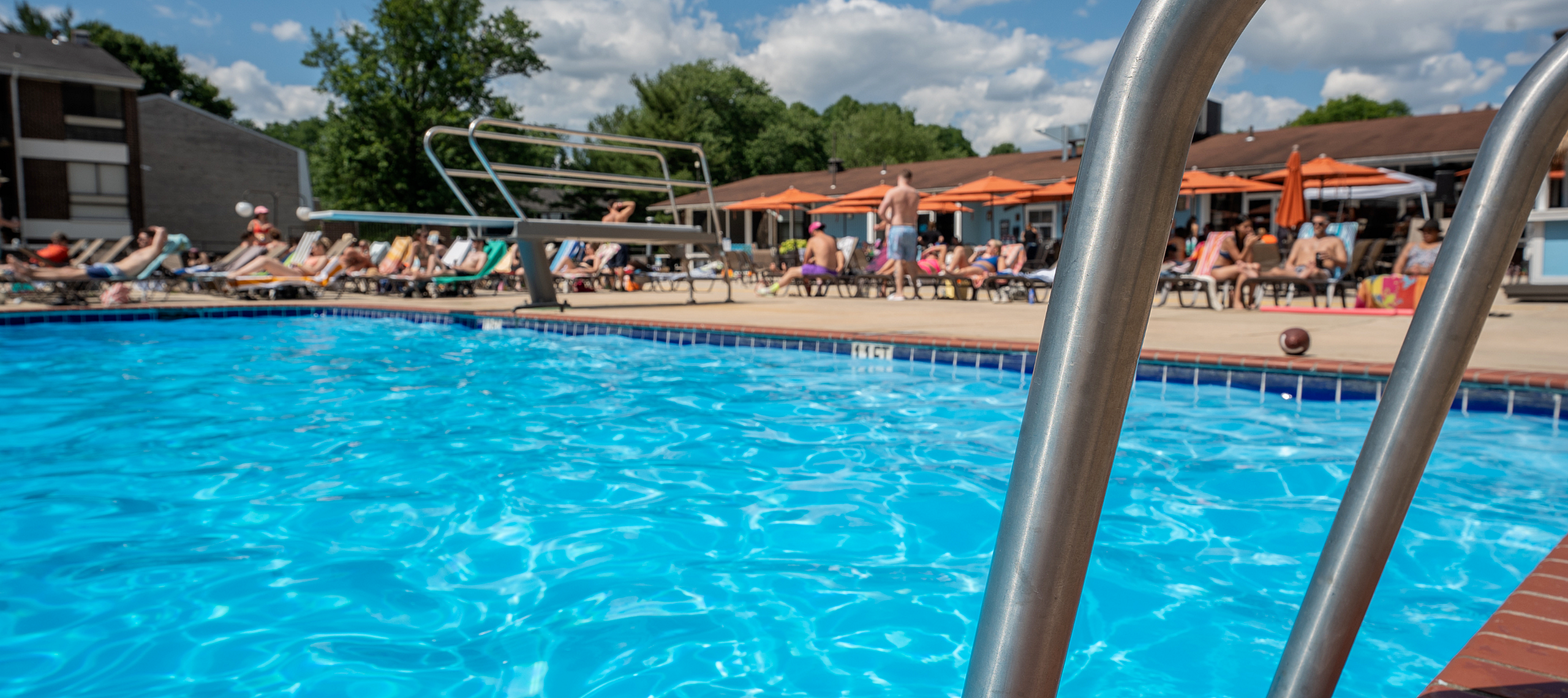 Vibrant poolside scene with clear blue water, surrounded by sun loungers and people relaxing under orange umbrellas on a sunny day.