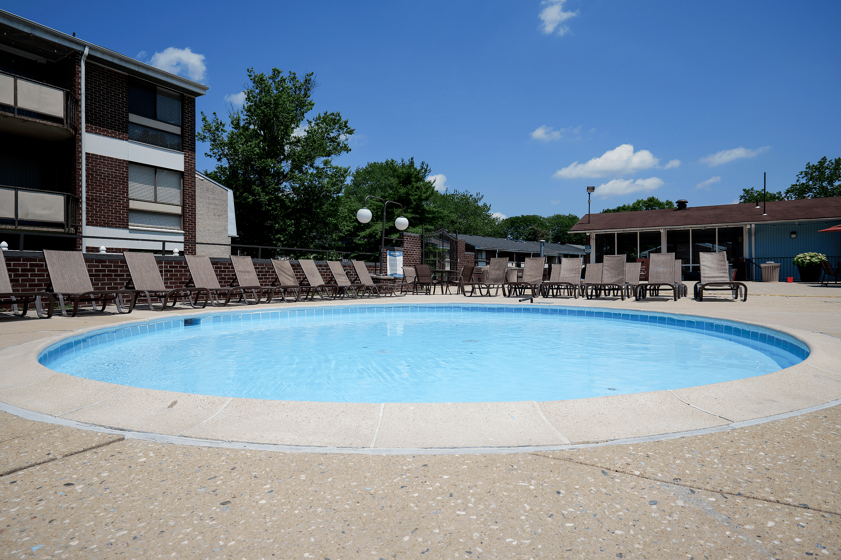 Charming apartment complex featuring a refreshing circular swimming pool surrounded by comfortable lounge chairs, set against a backdrop of lush trees and a clear blue sky.