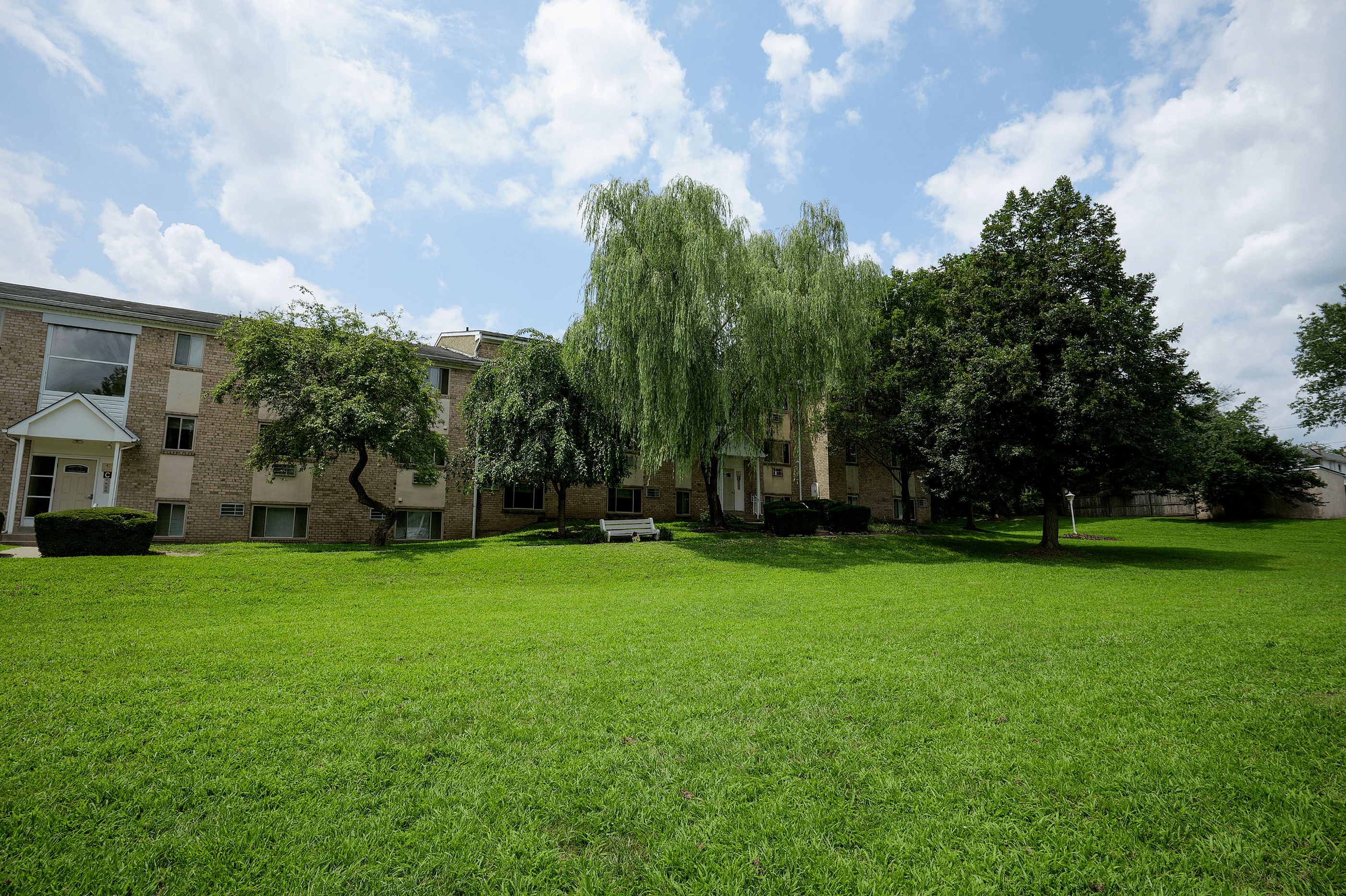 Charming apartment complex with lush green lawns and mature trees set against a bright blue sky.