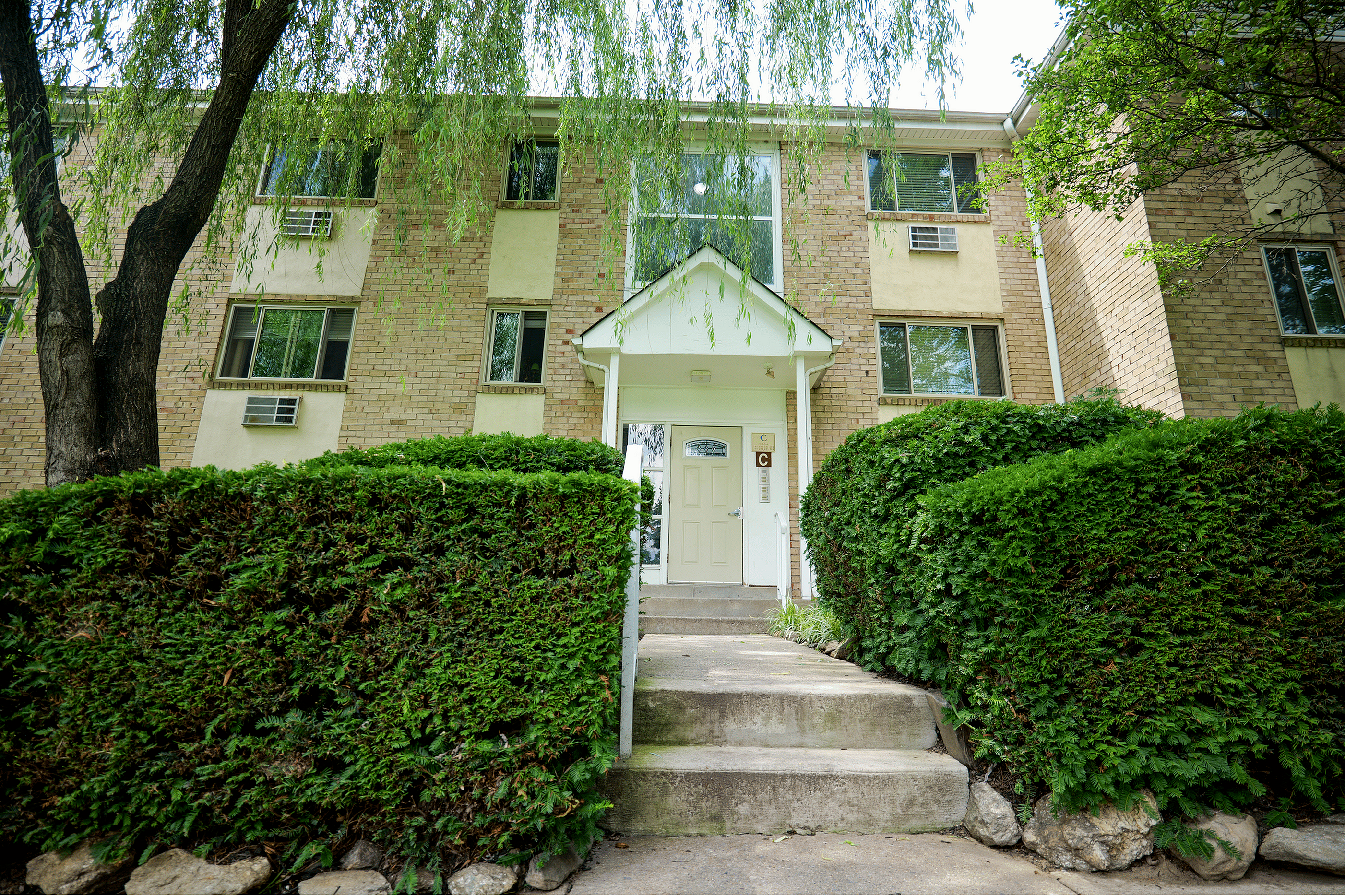 Inviting apartment entrance framed by lush greenery and classic brick facade, showcasing a tranquil residential oasis.