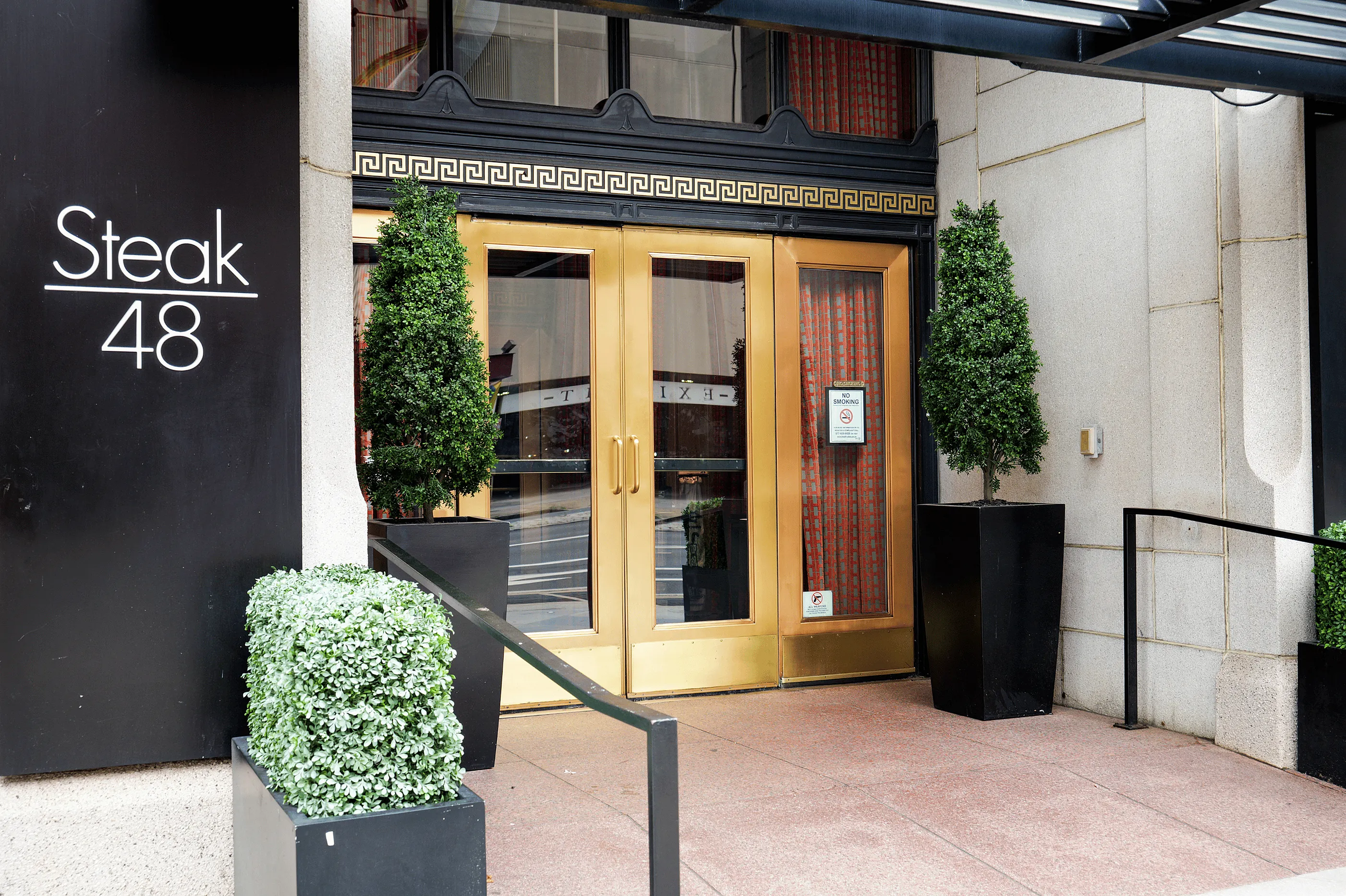 Elegant restaurant entrance with stylish gold doors and chic greenery, exuding sophistication and welcoming charm.