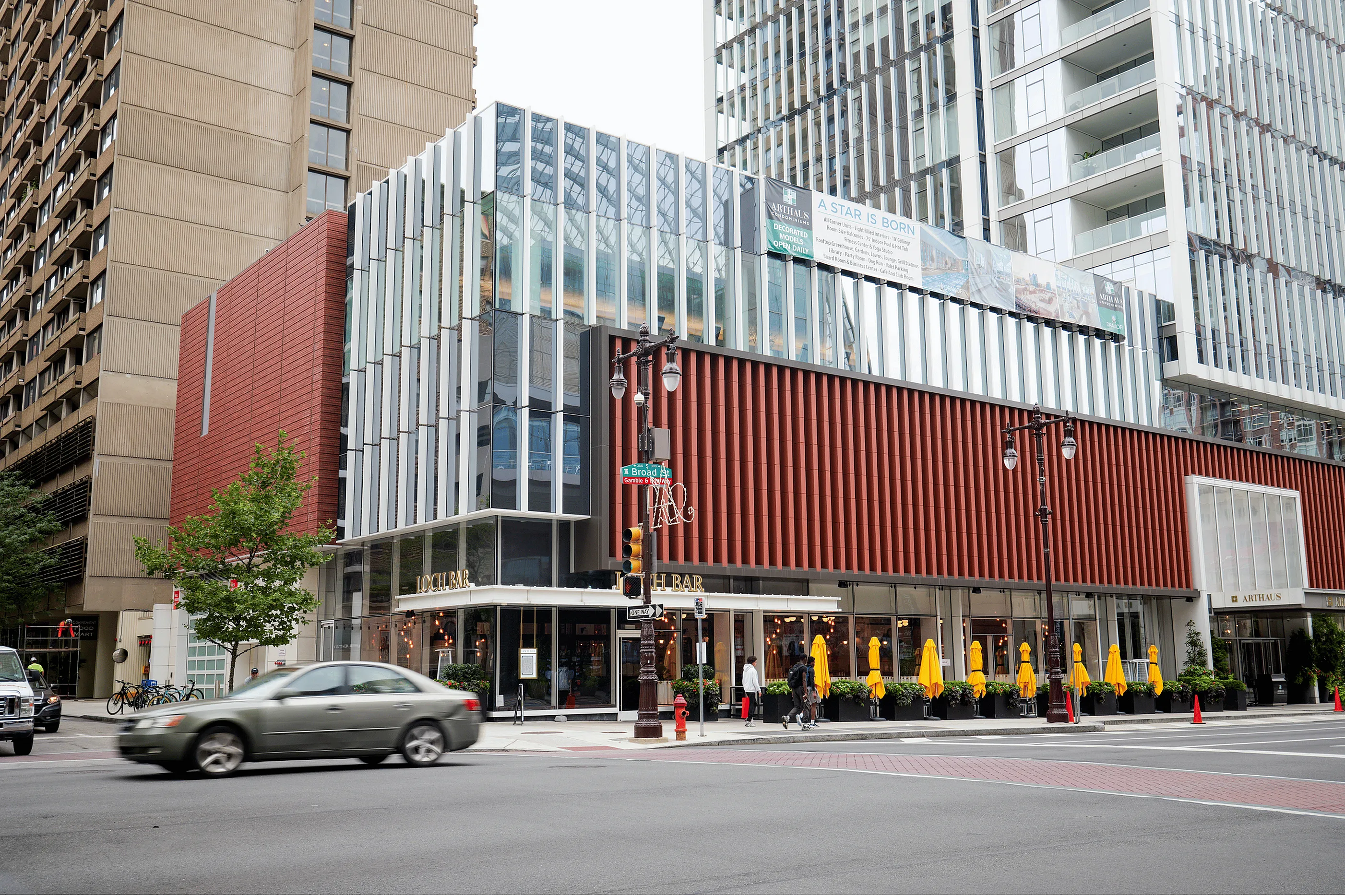 Modern urban architecture featuring a stylish multi-story building with sleek glass paneling and an inviting outdoor café with bright yellow umbrellas.