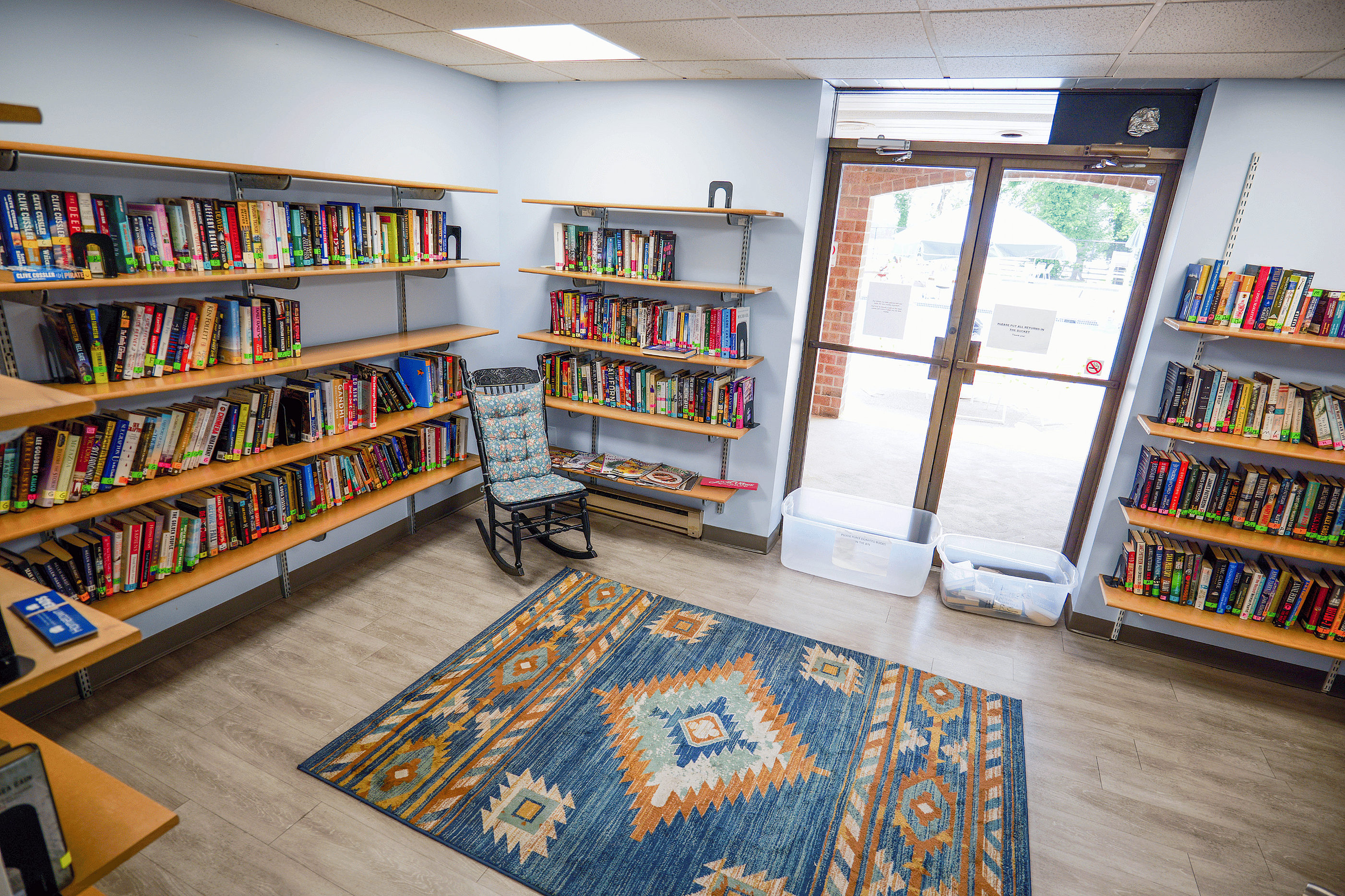 Cozy reading nook with bookshelves filled with colorful books, a comfy rocking chair, and an inviting patterned area rug next to large glass doors.