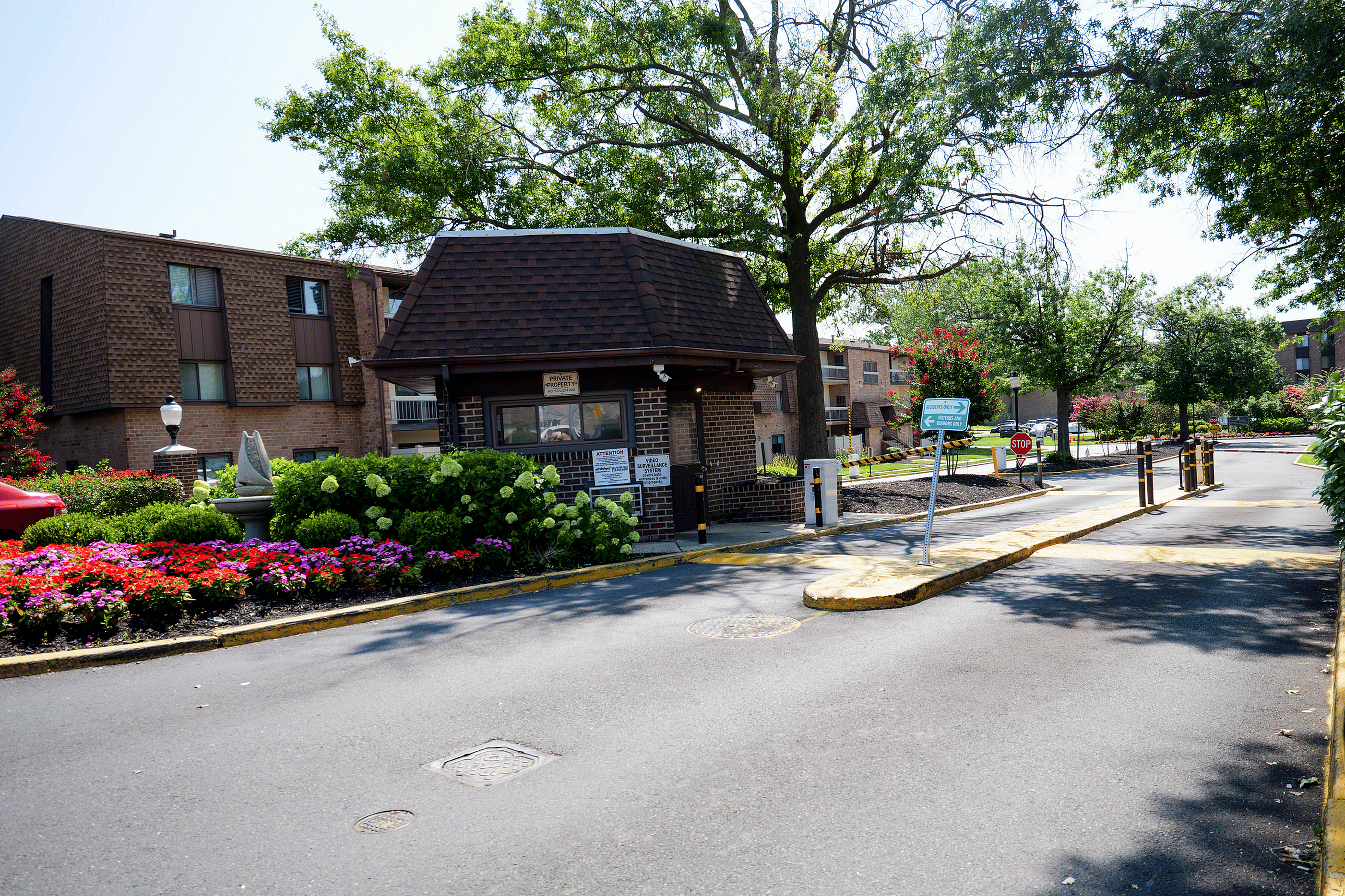 Charming gated community entrance with colorful flower beds and brick architecture, surrounded by lush greenery and mature trees.