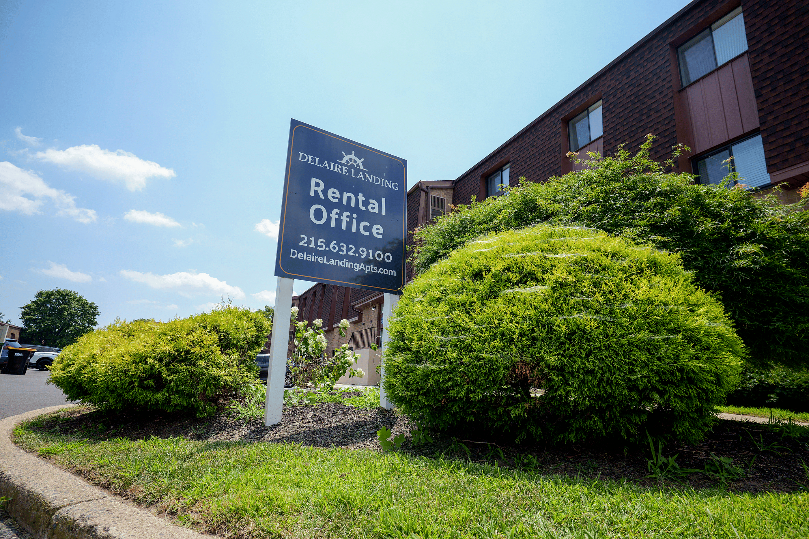 Welcoming rental office sign amidst lush greenery at Delaire Landing Apartments, basking in bright sunlight.
