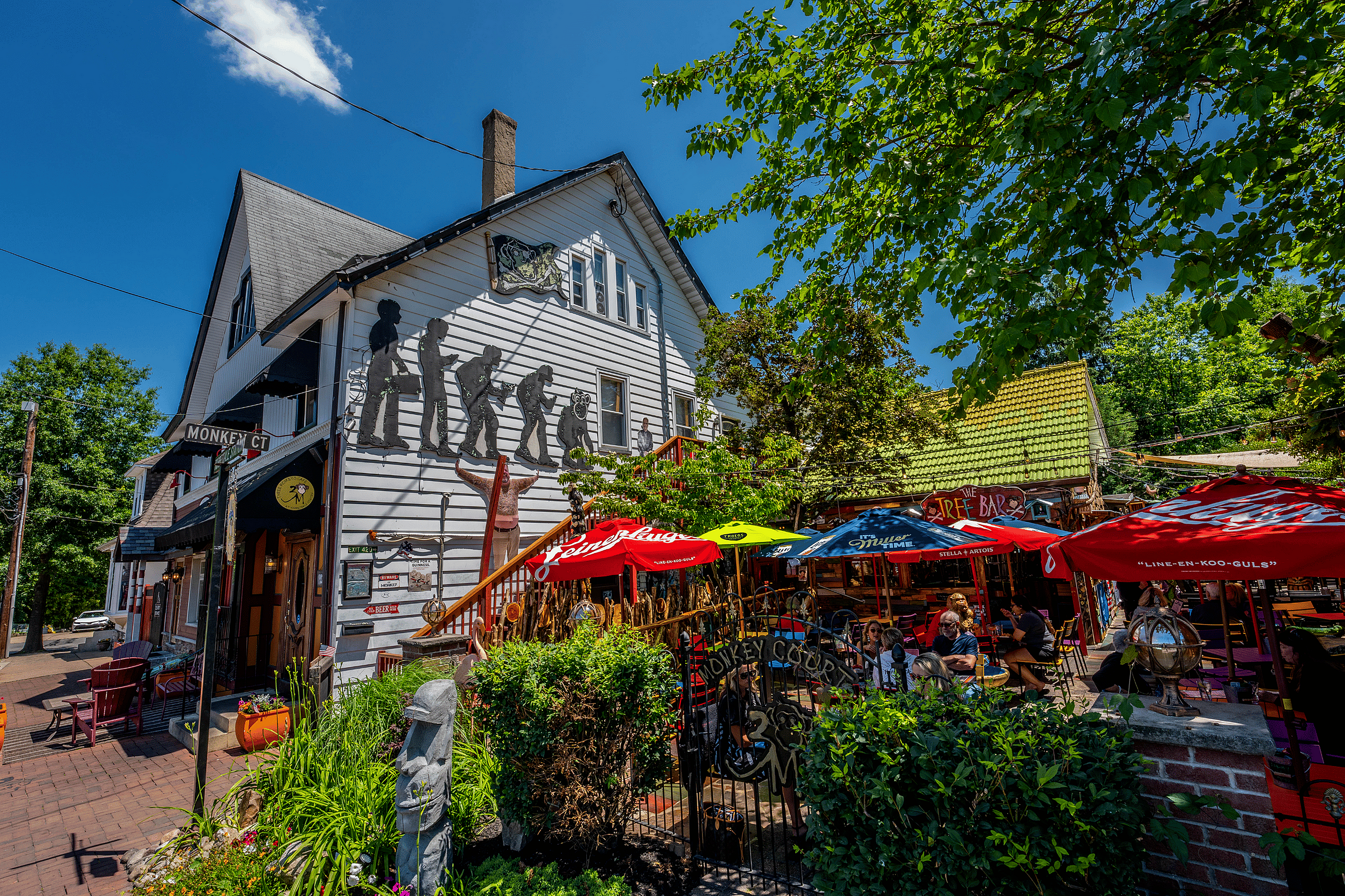 Charming two-story building with artistic decorations and colorful outdoor seating under vibrant umbrellas, nestled in lush greenery on a sunny day.
