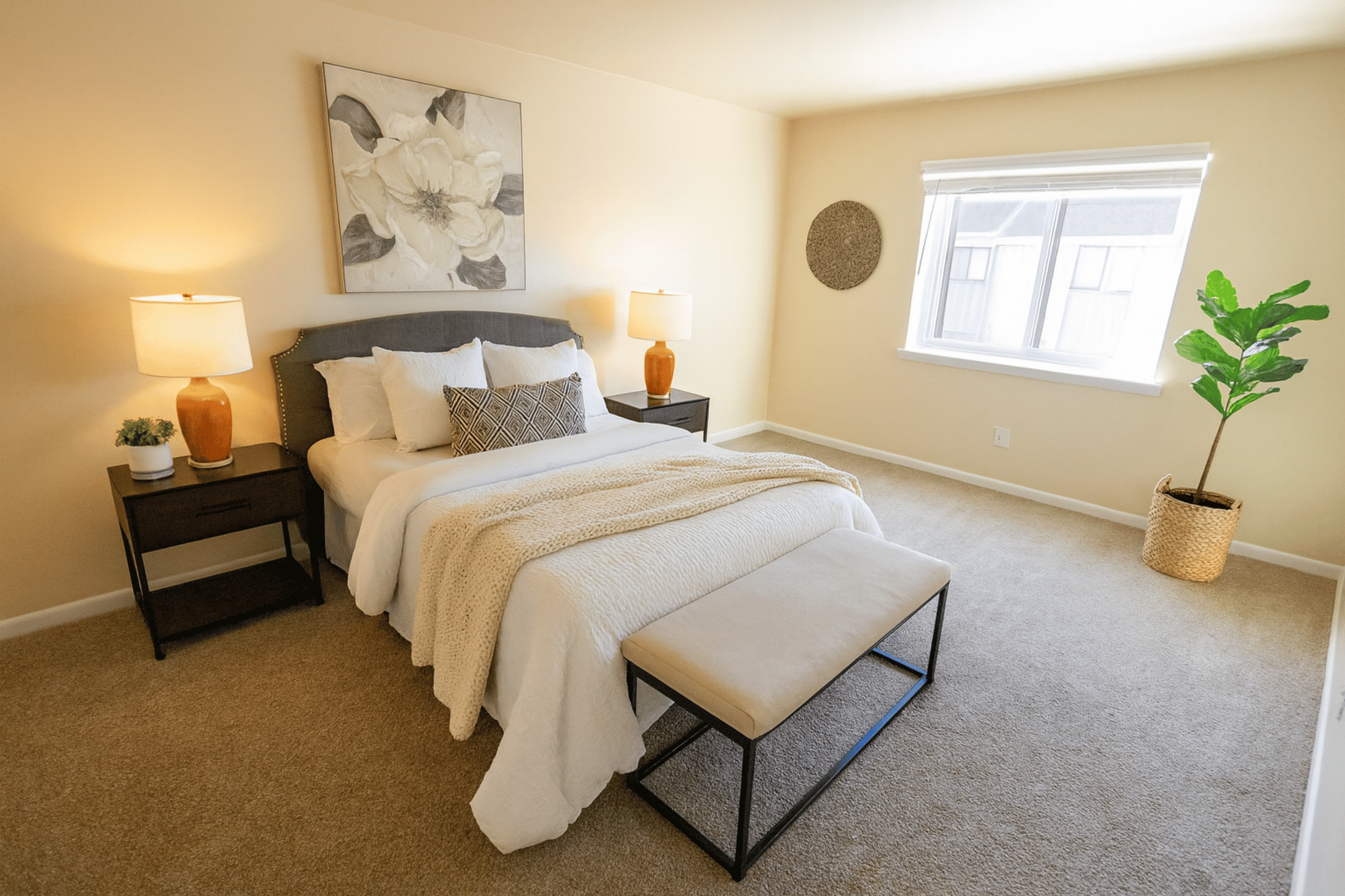 Inviting bedroom with warm lighting, featuring a plush bed with cozy textiles, elegant nightstands, and a large window providing natural light. Minimalistic decor includes a stylish plant and floral artwork, creating a serene ambiance.