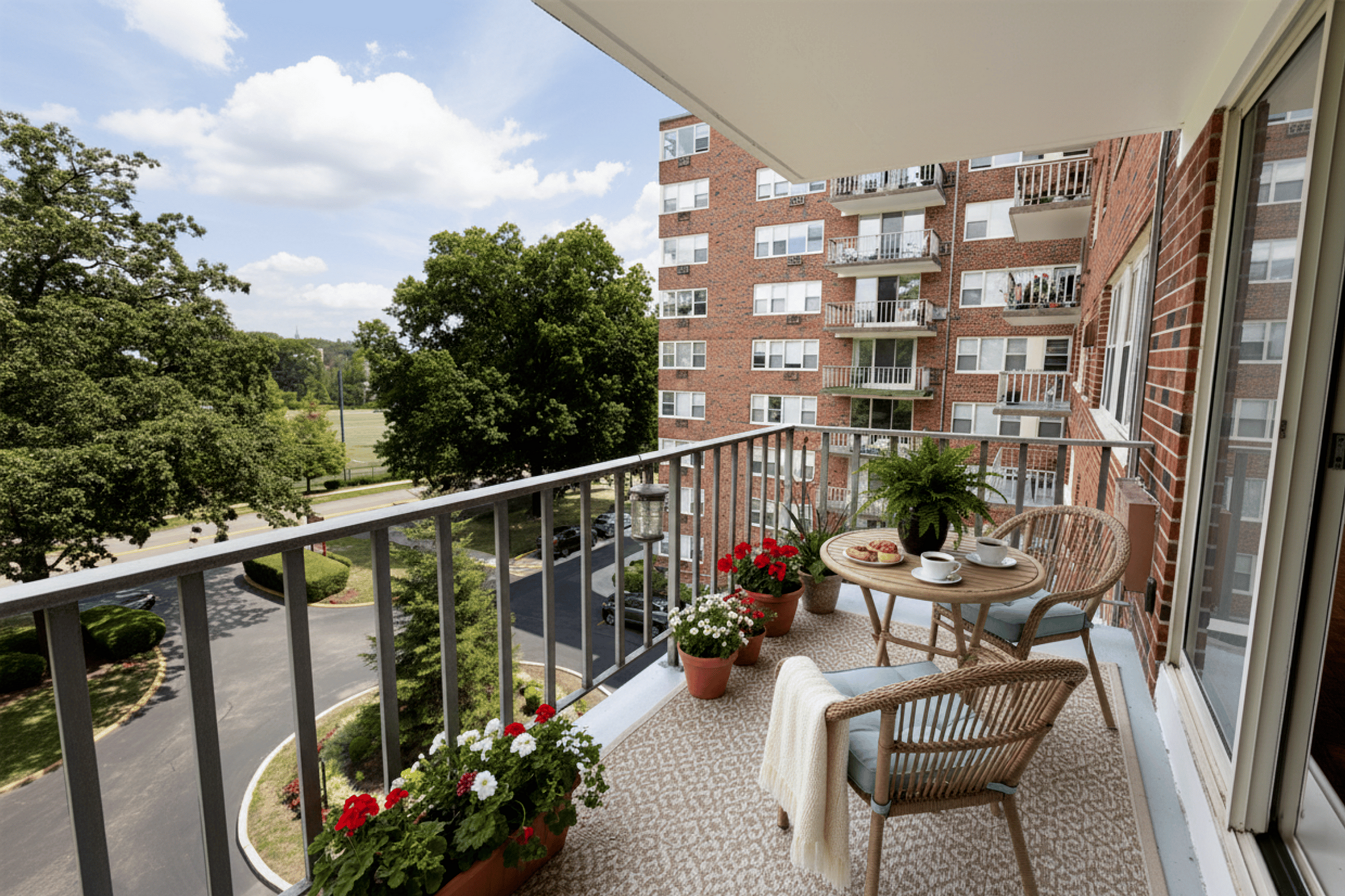Charming apartment balcony with cozy outdoor seating, vibrant flowers, and scenic views of lush trees and neighboring buildings.