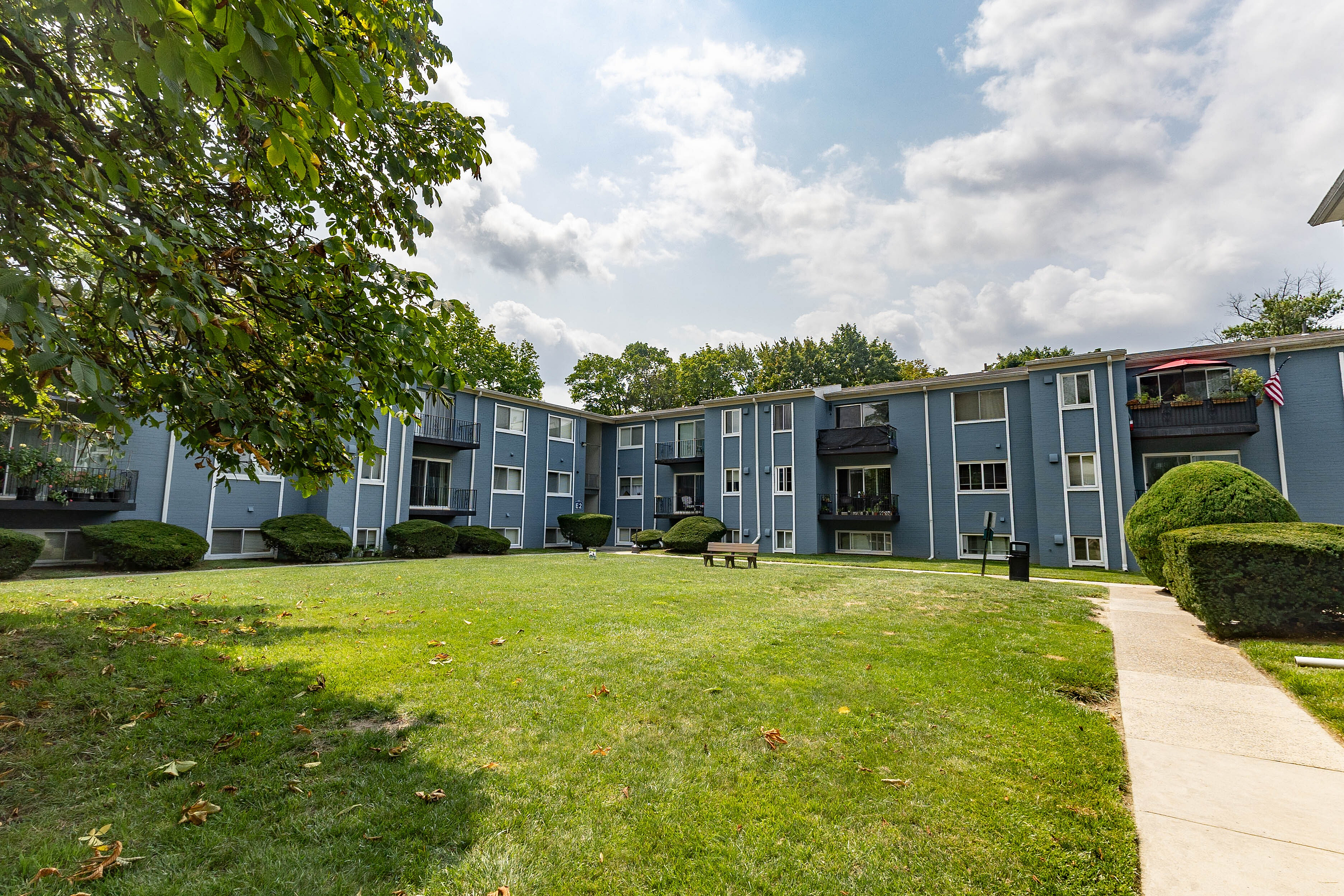 Charming apartment complex with a modern blue facade, set amidst lush green lawns and mature trees under a picturesque sky.