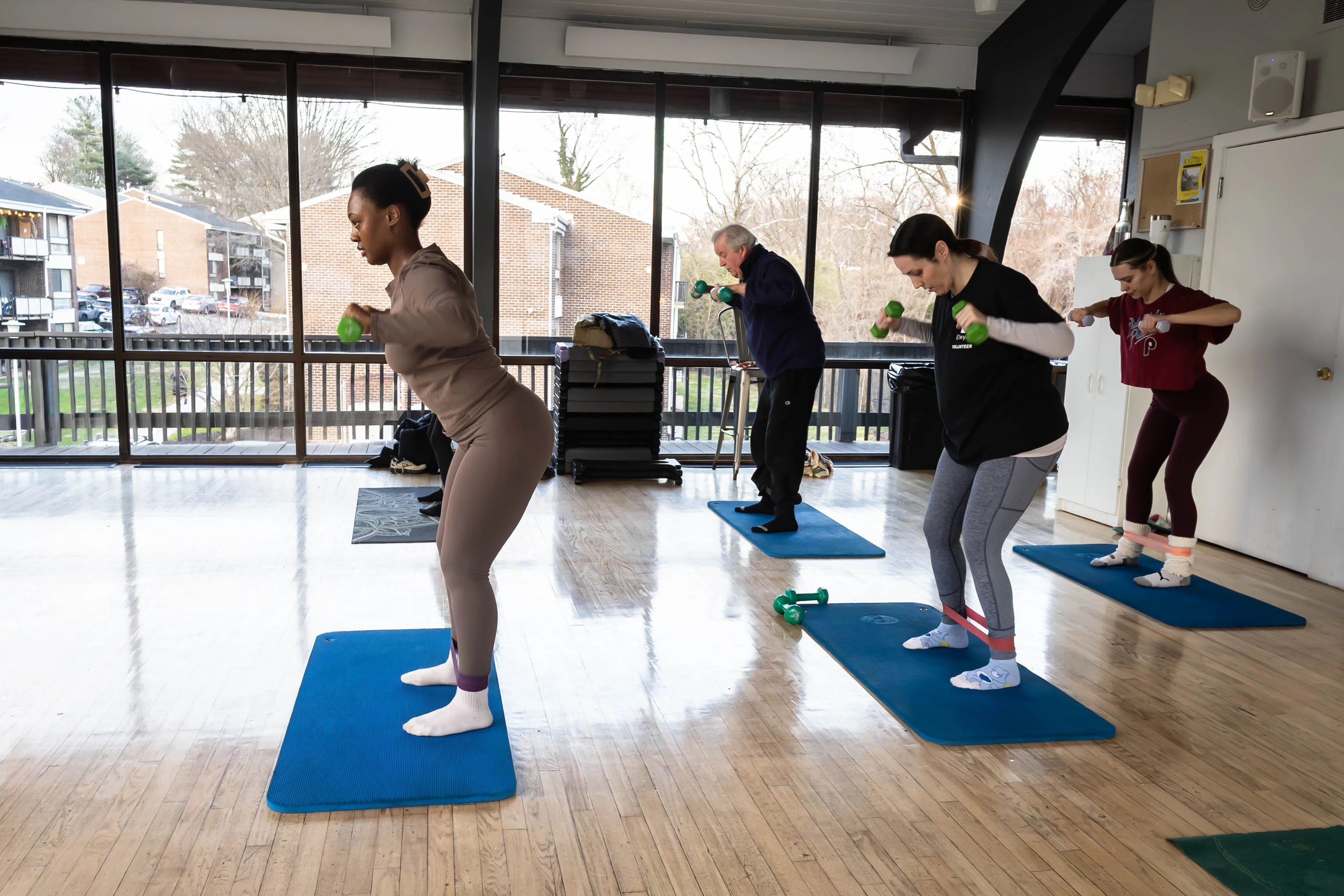 Bright fitness studio with large windows, featuring a diverse group of people engaging in energetic exercise routines on yoga mats with dumbbells.