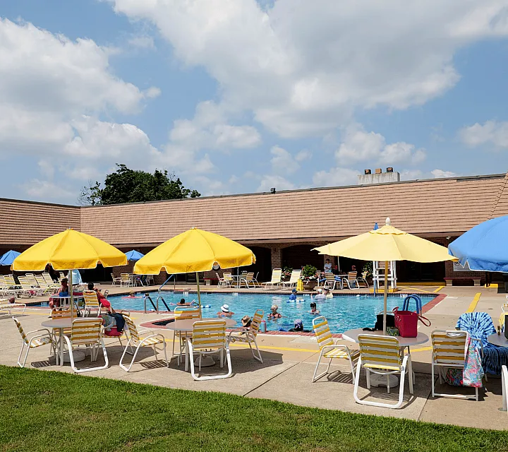 Inviting apartment complex pool area with vibrant yellow and blue umbrellas, surrounded by comfortable lounge chairs and lush green lawns.