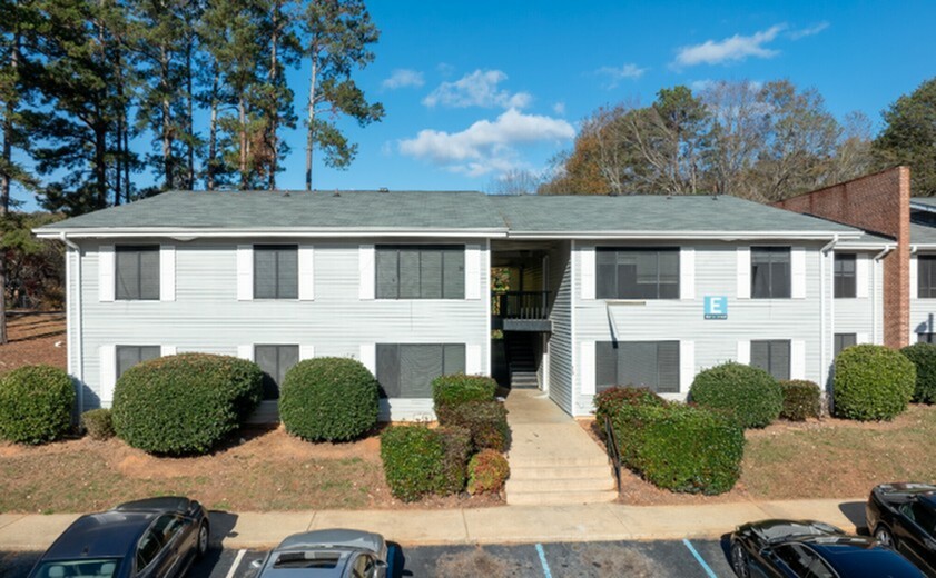 Charming multi-unit apartment building with well-maintained landscaping, convenient parking, and surrounded by lush trees under a clear blue sky.
